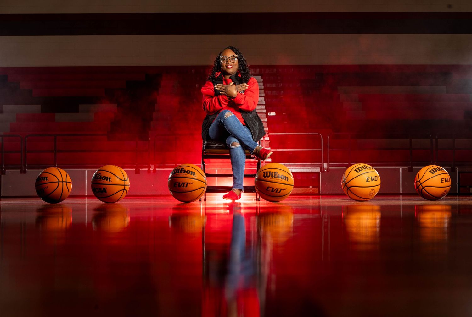 Woman seated on a chair surrounded by basketballs on a court with dramatic red lighting.