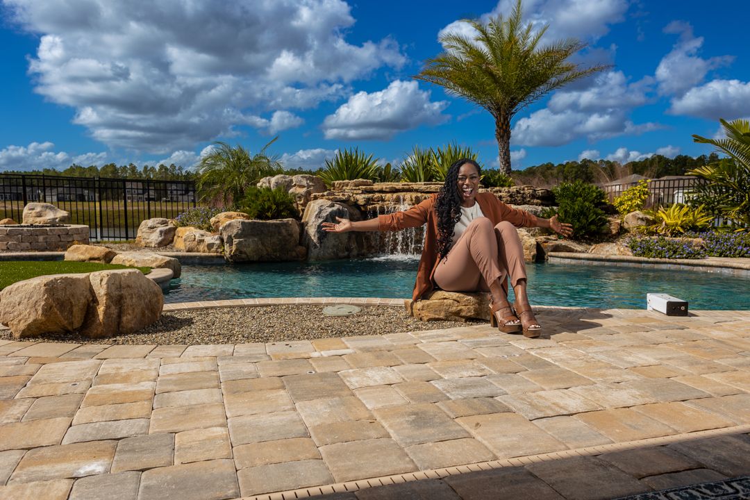 Woman posing beside a luxury pool with a waterfall feature and palm trees under blue skies.