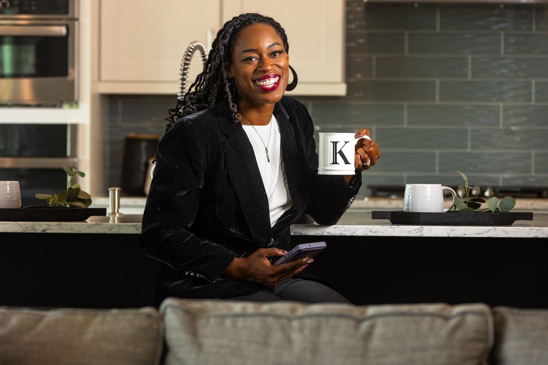 Stylish woman in black blazer holding mug and phone smiling in modern kitchen.