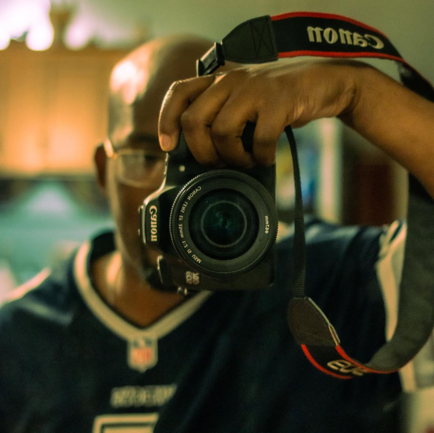 Photographer holding a Canon DSLR camera up to their face wearing an NFL jersey.