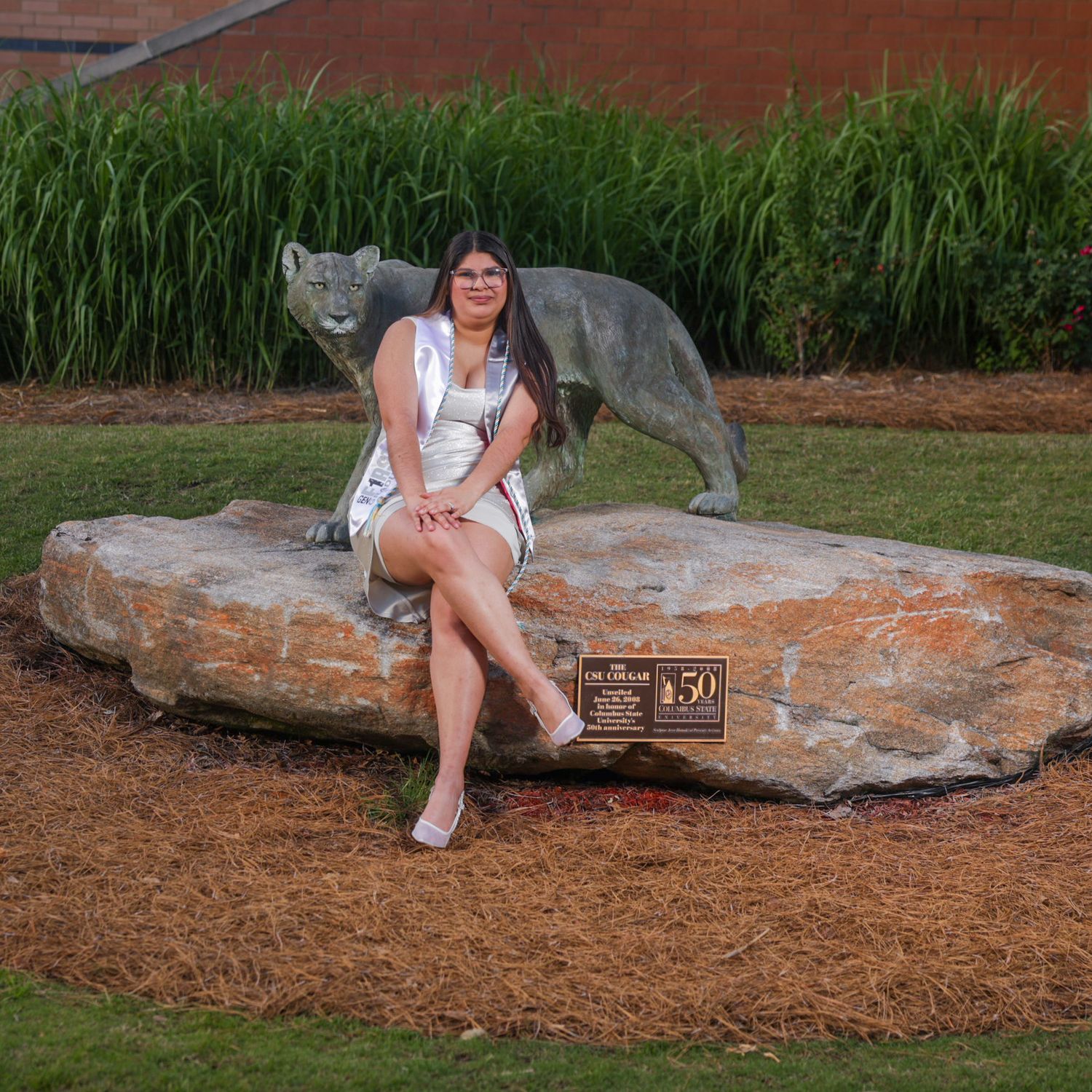 Young woman in white dress sitting on a rock beside a cougar statue on a college campus.