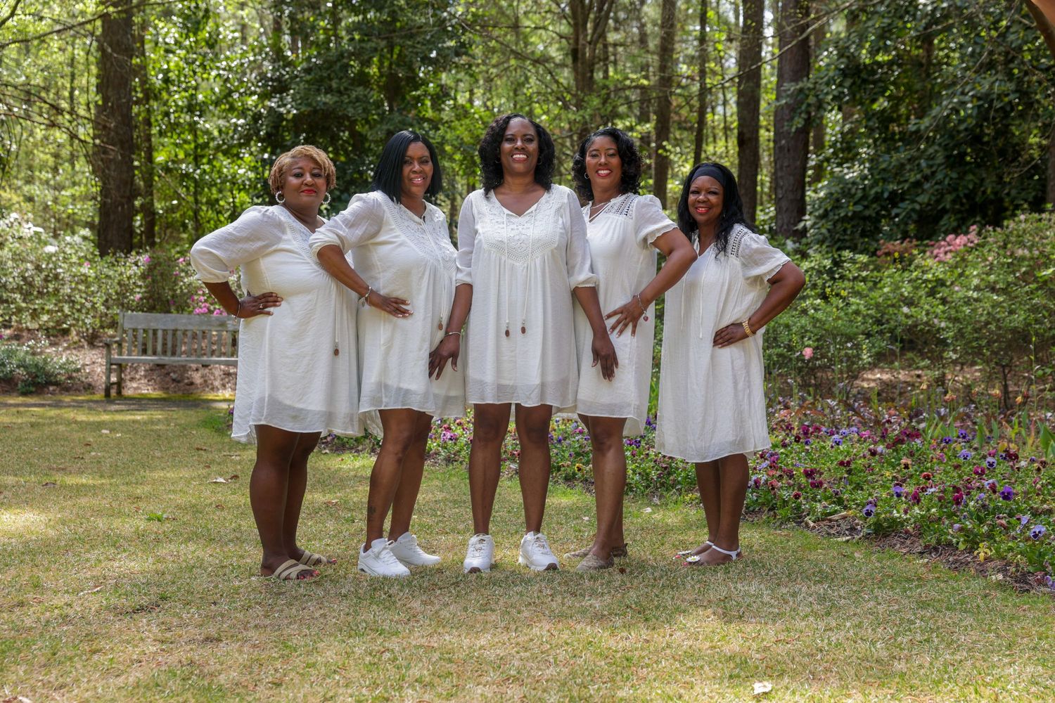 Five women in matching white dresses posing together in a lush green garden.