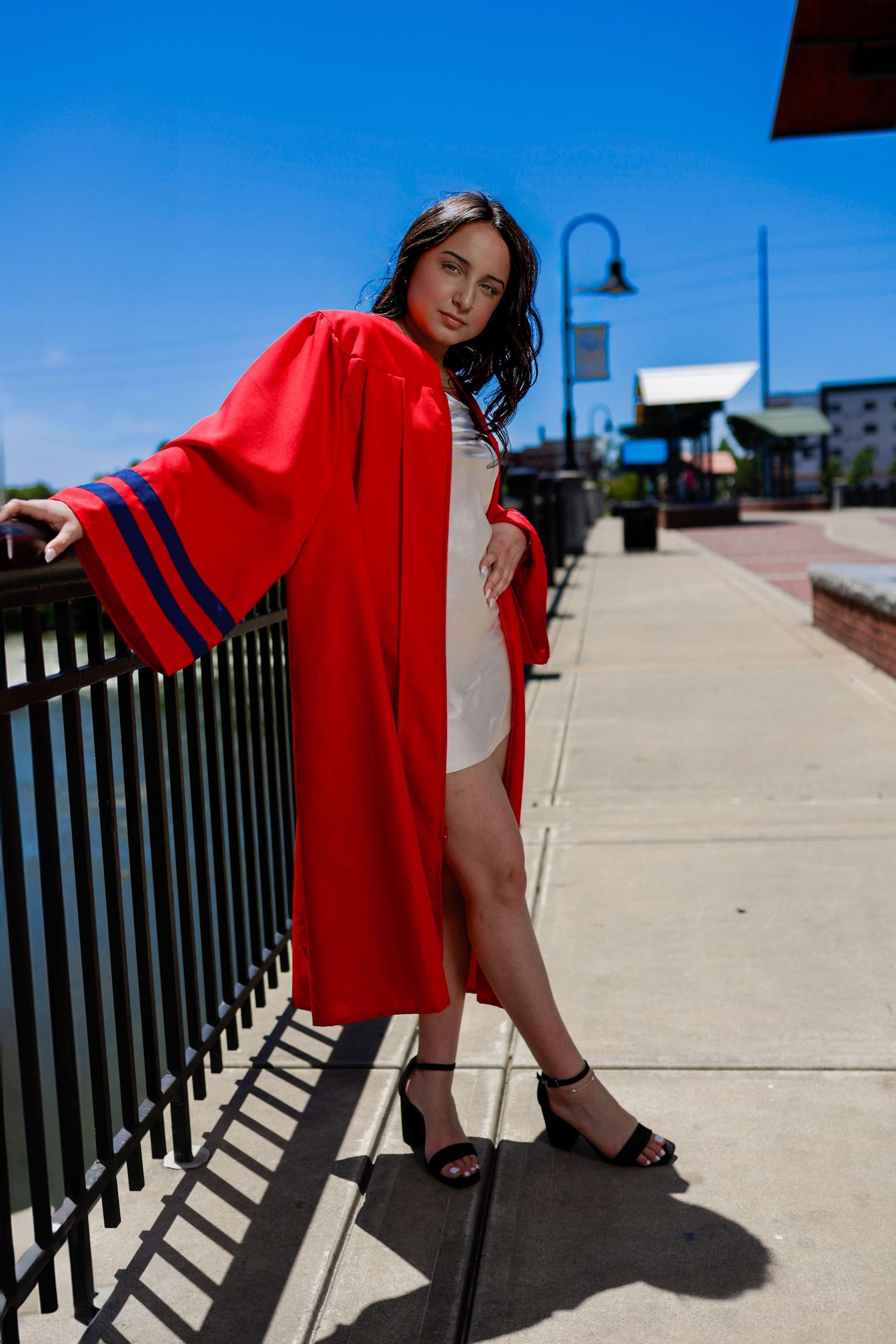 Young woman in red graduation gown posing by iron fence on sunny day, wearing black heels and white dress.