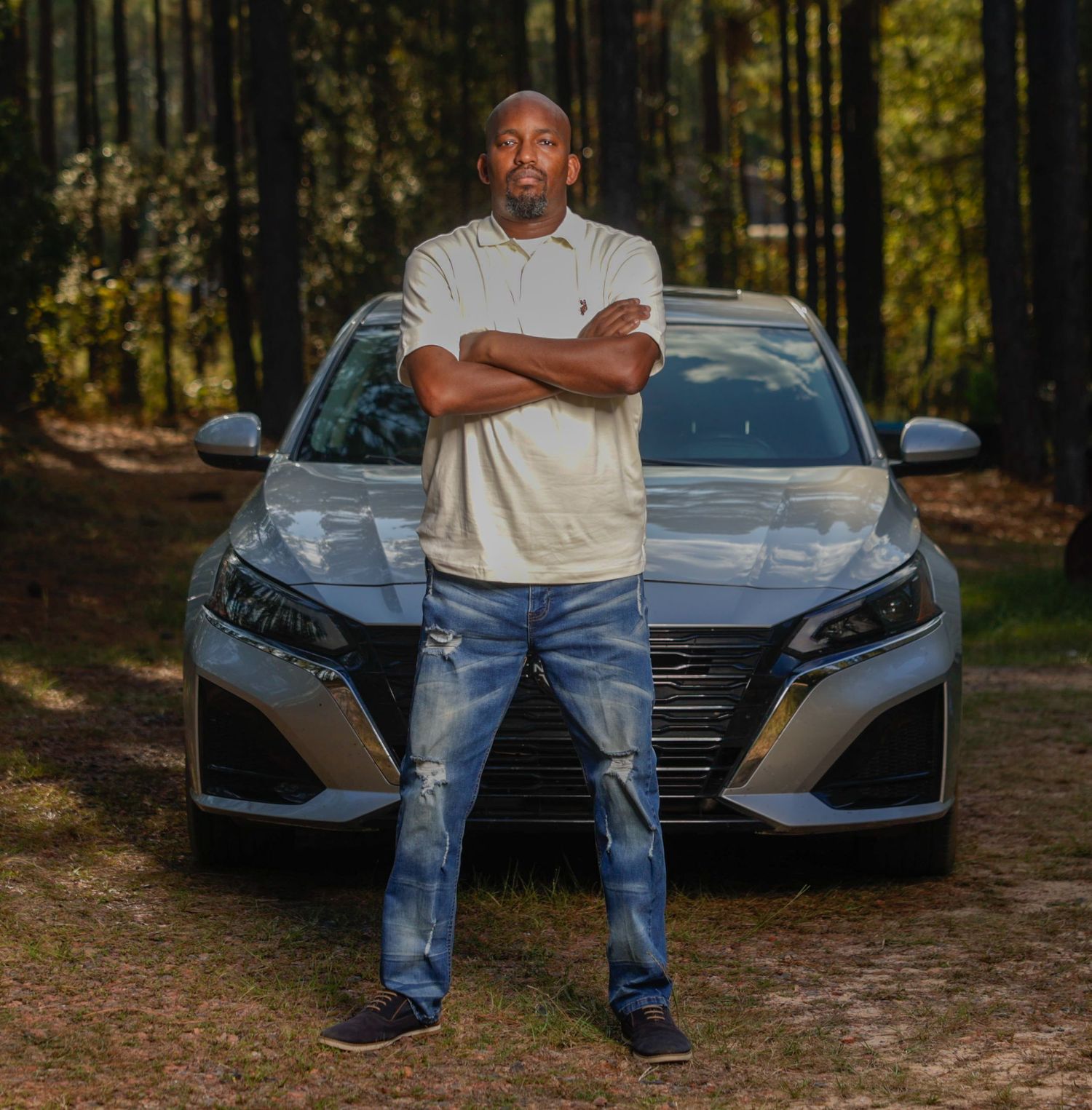Man in beige polo and jeans standing confidently in front of a silver sedan in a forest.