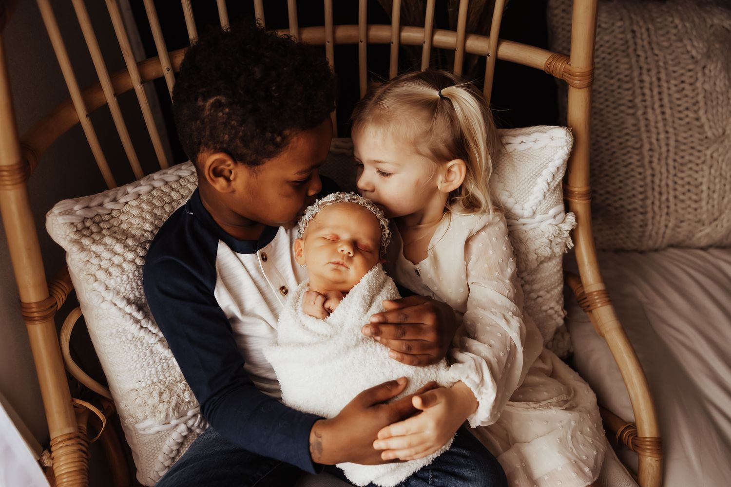 Two children lovingly hold a swaddled newborn baby while sitting together in a rattan chair.
