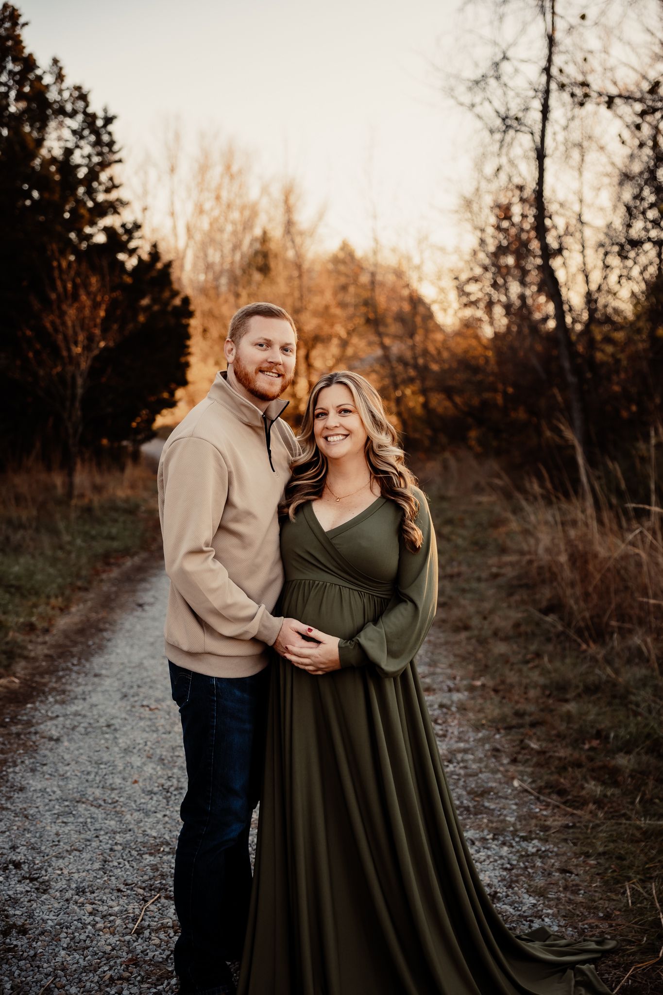 A couple poses on a gravel path at golden hour, the woman in an olive maternity gown cradling her bump.