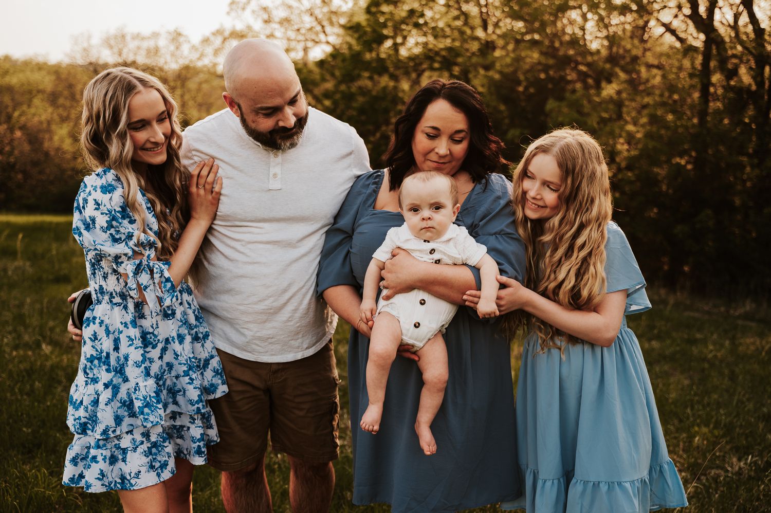 Happy family of five posing outdoors at golden hour, parents holding baby boy surrounded by two blonde girls in blue dresses.