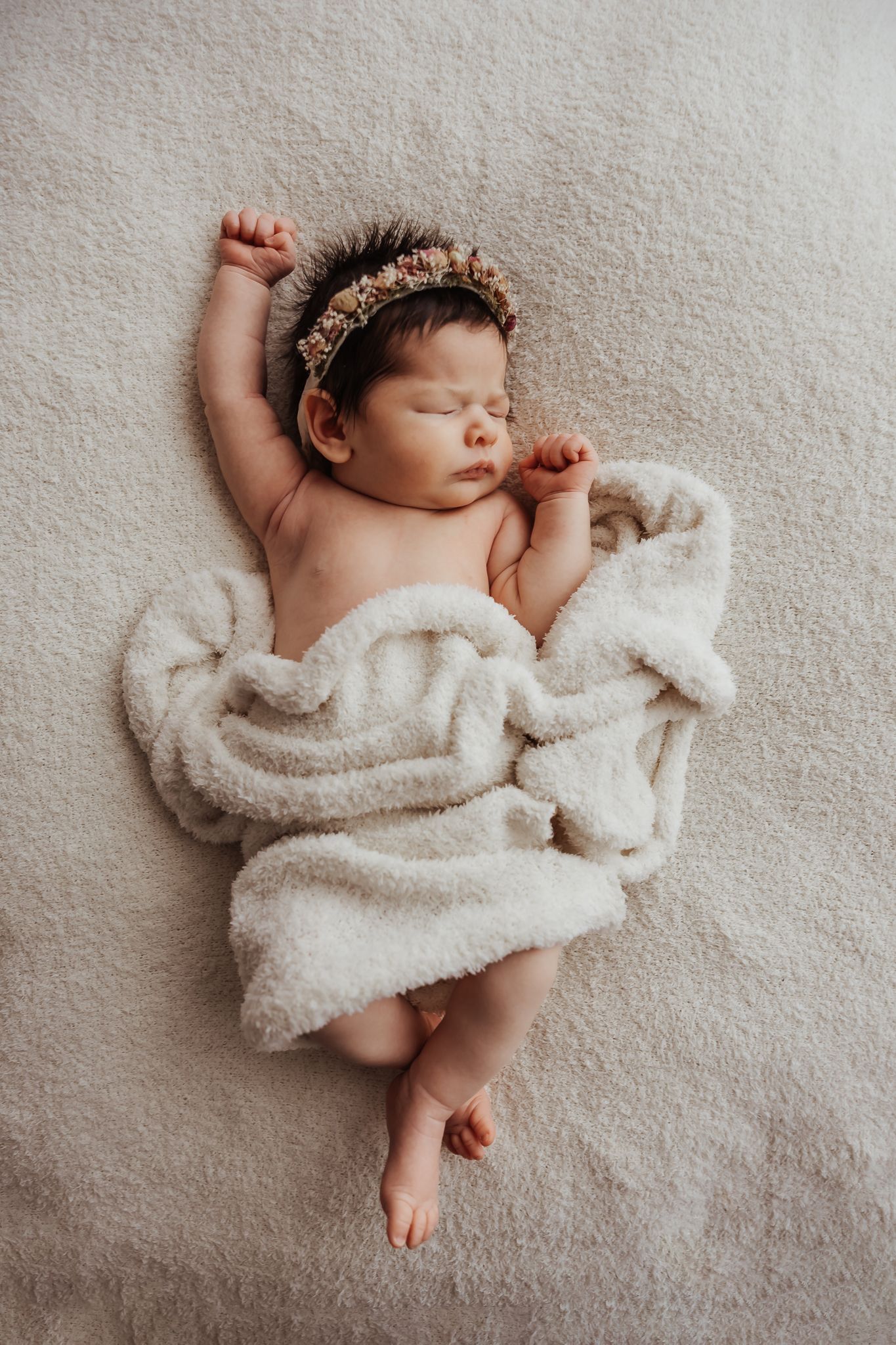 Sleeping newborn baby girl wearing a floral crown, wrapped in a soft white blanket on a cream background.