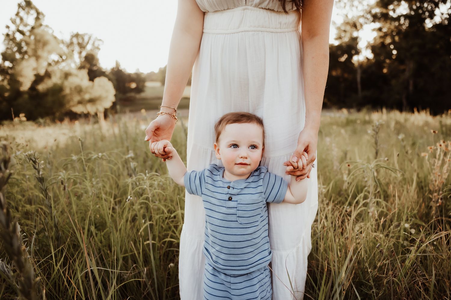 A mother in a white dress helps her baby boy take first steps in a sunny green meadow.