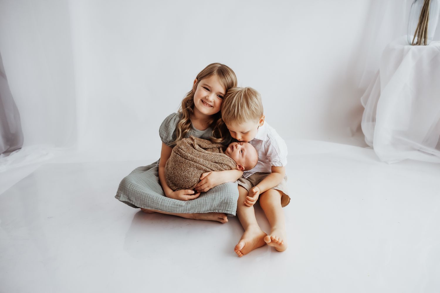 Two siblings lovingly hold their newborn baby in a bright white studio during a family portrait session.