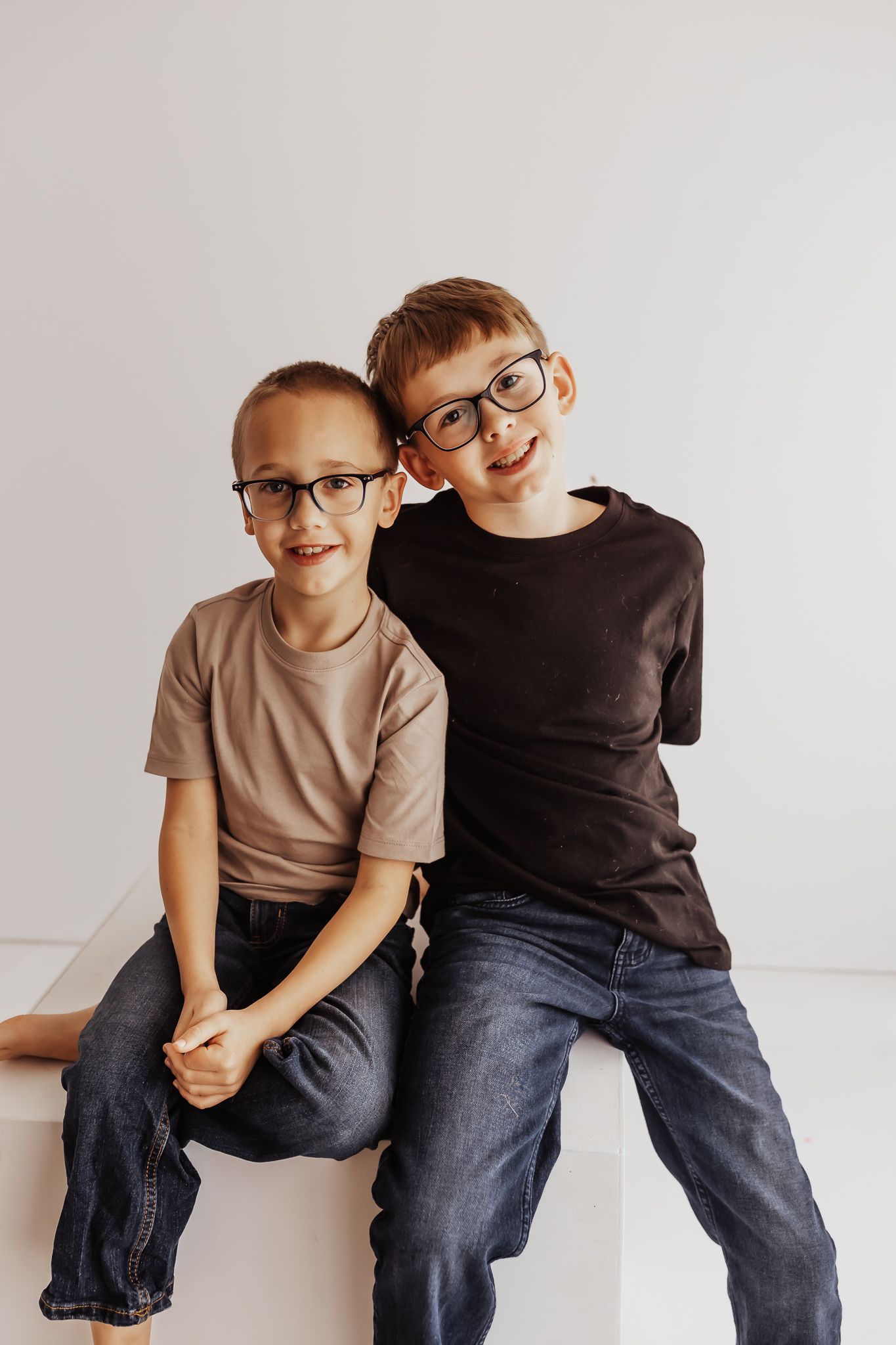 Two young boys wearing glasses and casual clothes sitting together smiling in a bright studio.