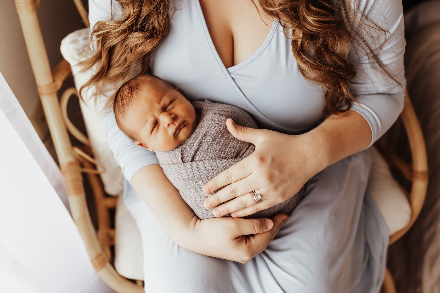 Mother holding sleeping newborn baby wrapped in gray swaddle while sitting in rattan chair.