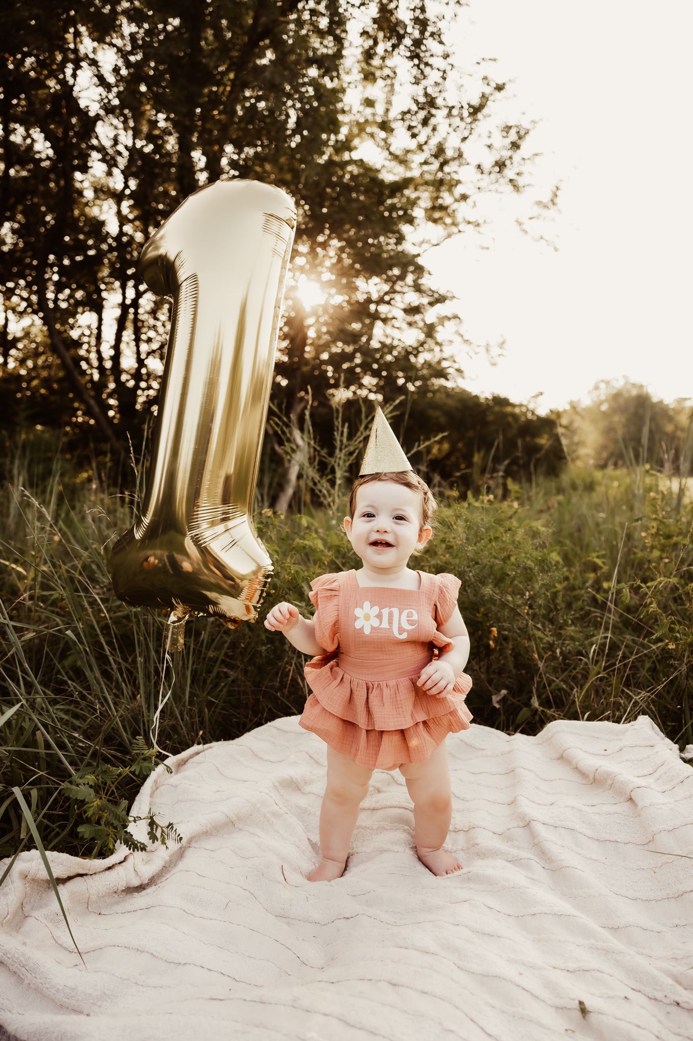 Happy baby girl standing with gold number one balloon and party hat celebrating first birthday.