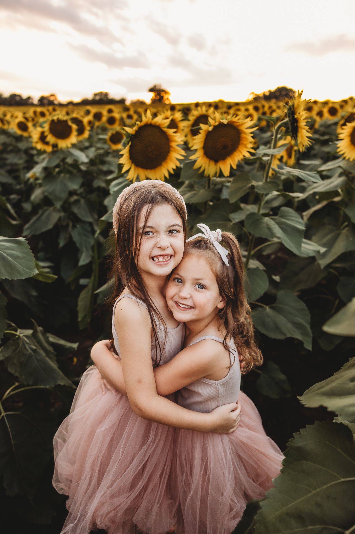 Two young sisters hugging and smiling in pink tulle dresses in a sunflower field at sunset.
