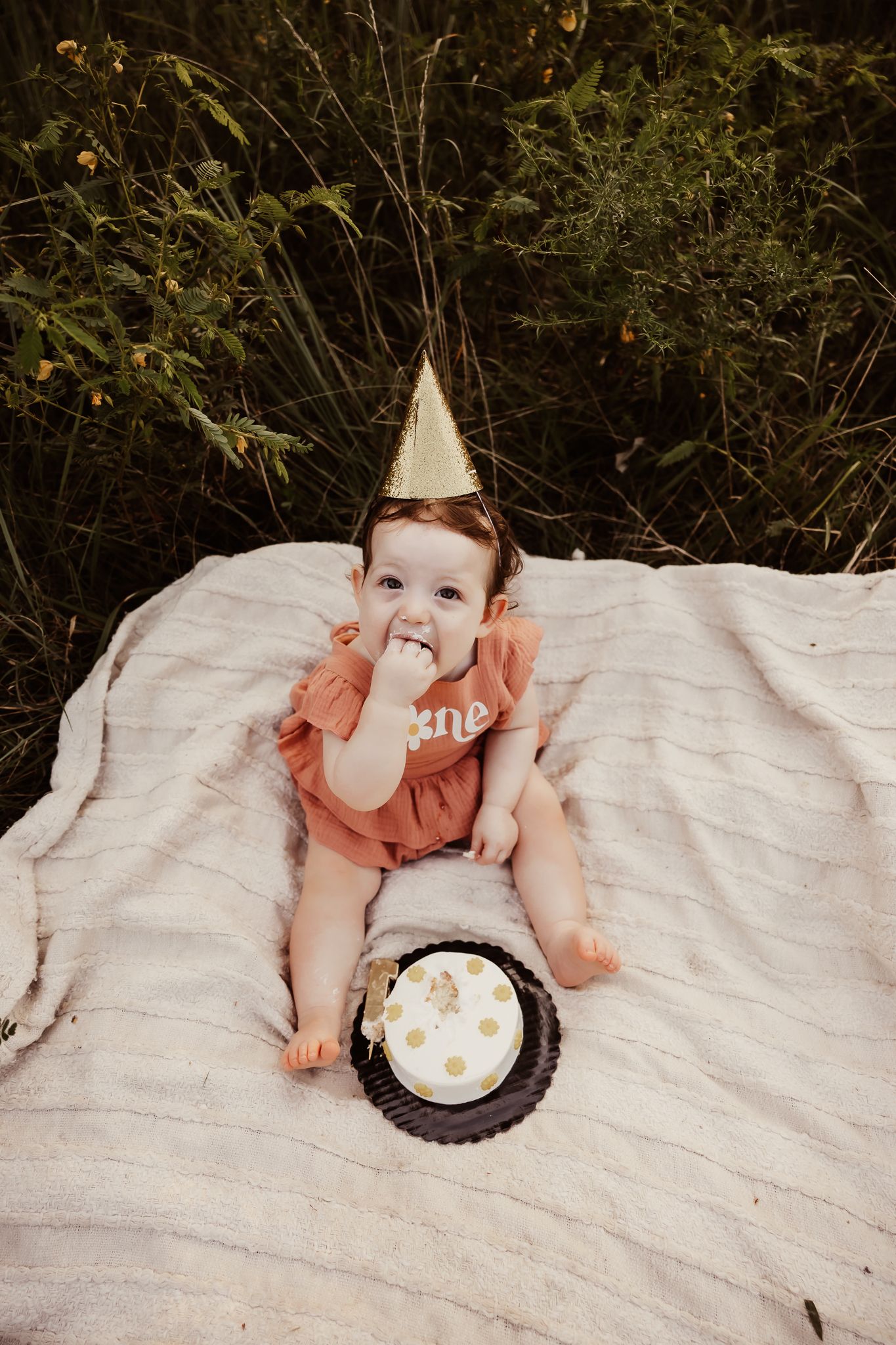 Baby girl in rust romper and gold party hat eating first birthday cake smash outdoors.