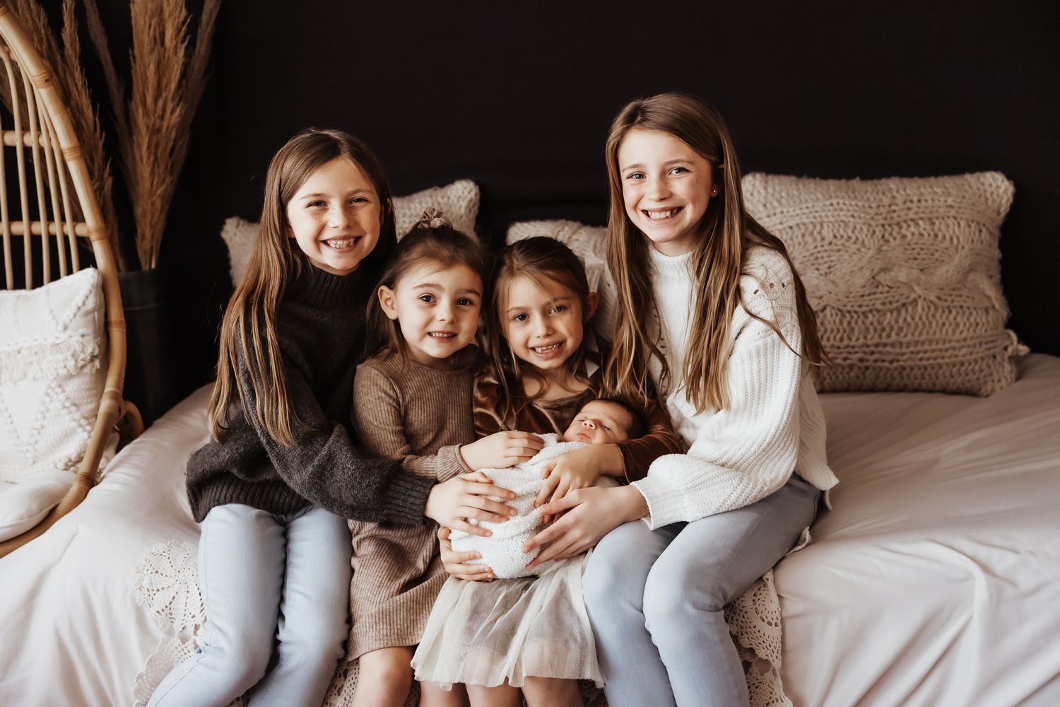 Four smiling girls sitting on a bed, with the oldest holding a newborn baby, in a cozy studio setting.