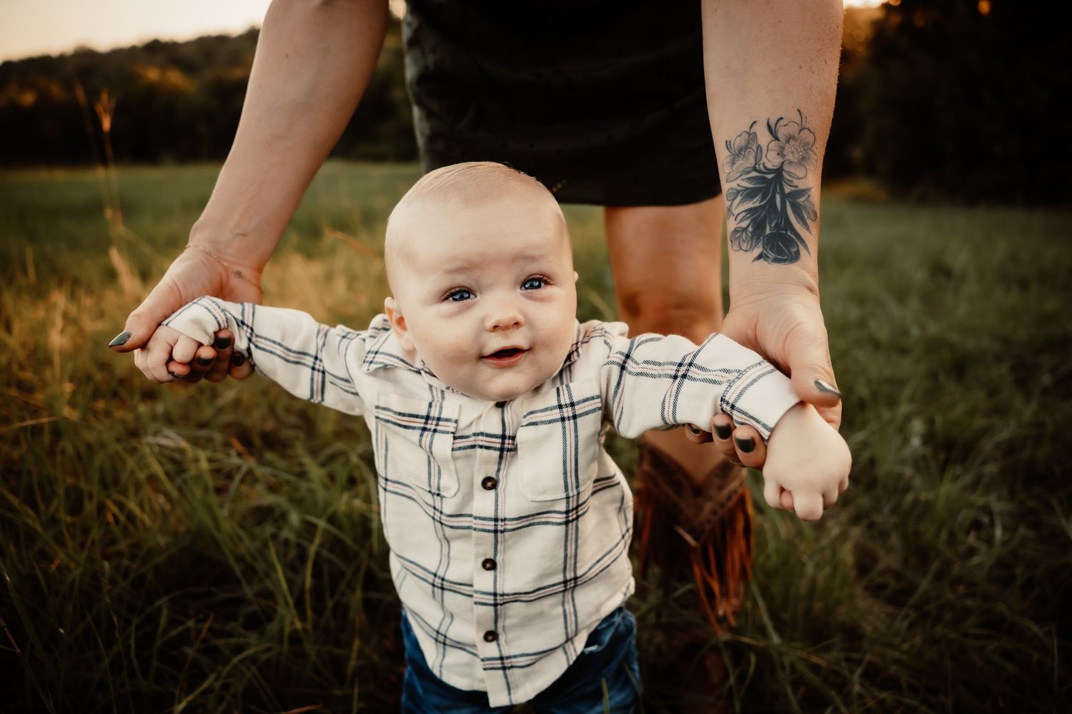 A smiling baby in a plaid shirt stands in a sunny field, supported by an adult with a tattoo on their leg.