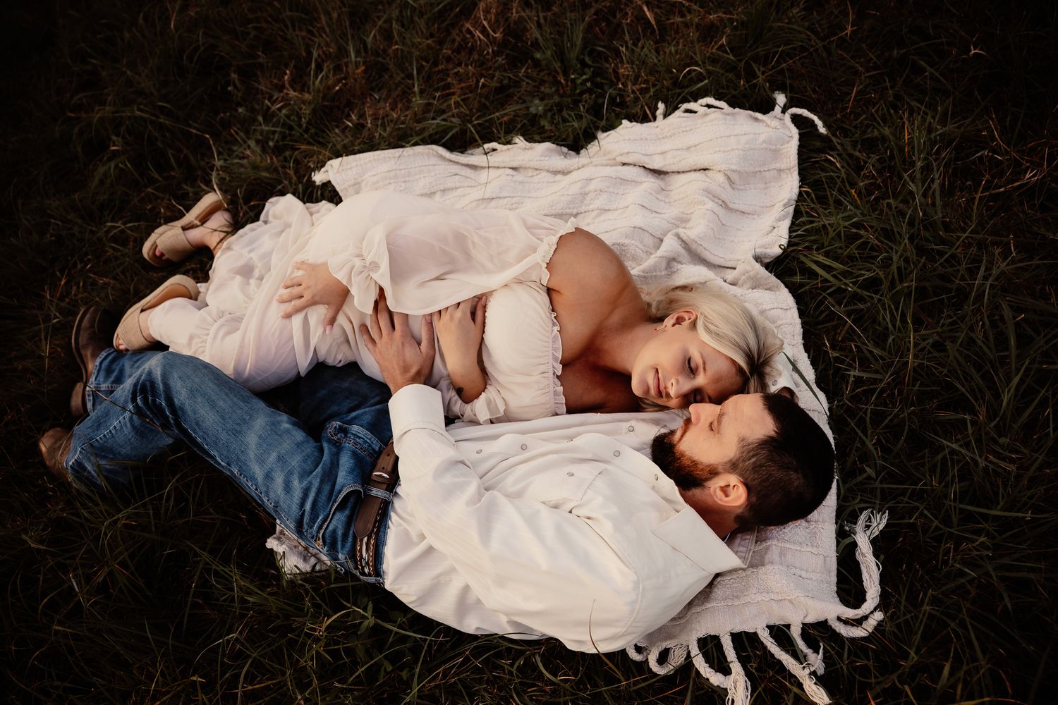 Pregnant woman and child lying on white blanket in grass during outdoor maternity photo session.