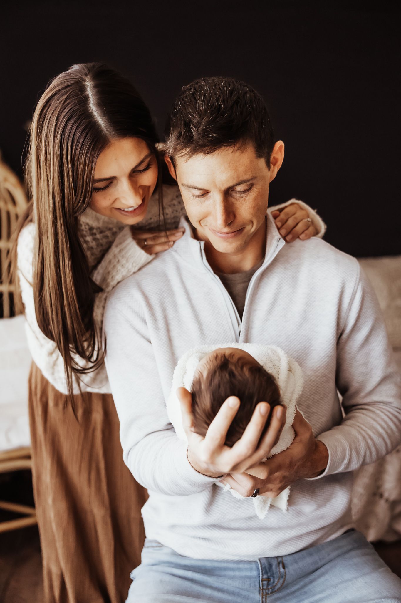 Young parents lovingly gaze at their newborn baby, father cradling infant while mother rests hand on his shoulder.