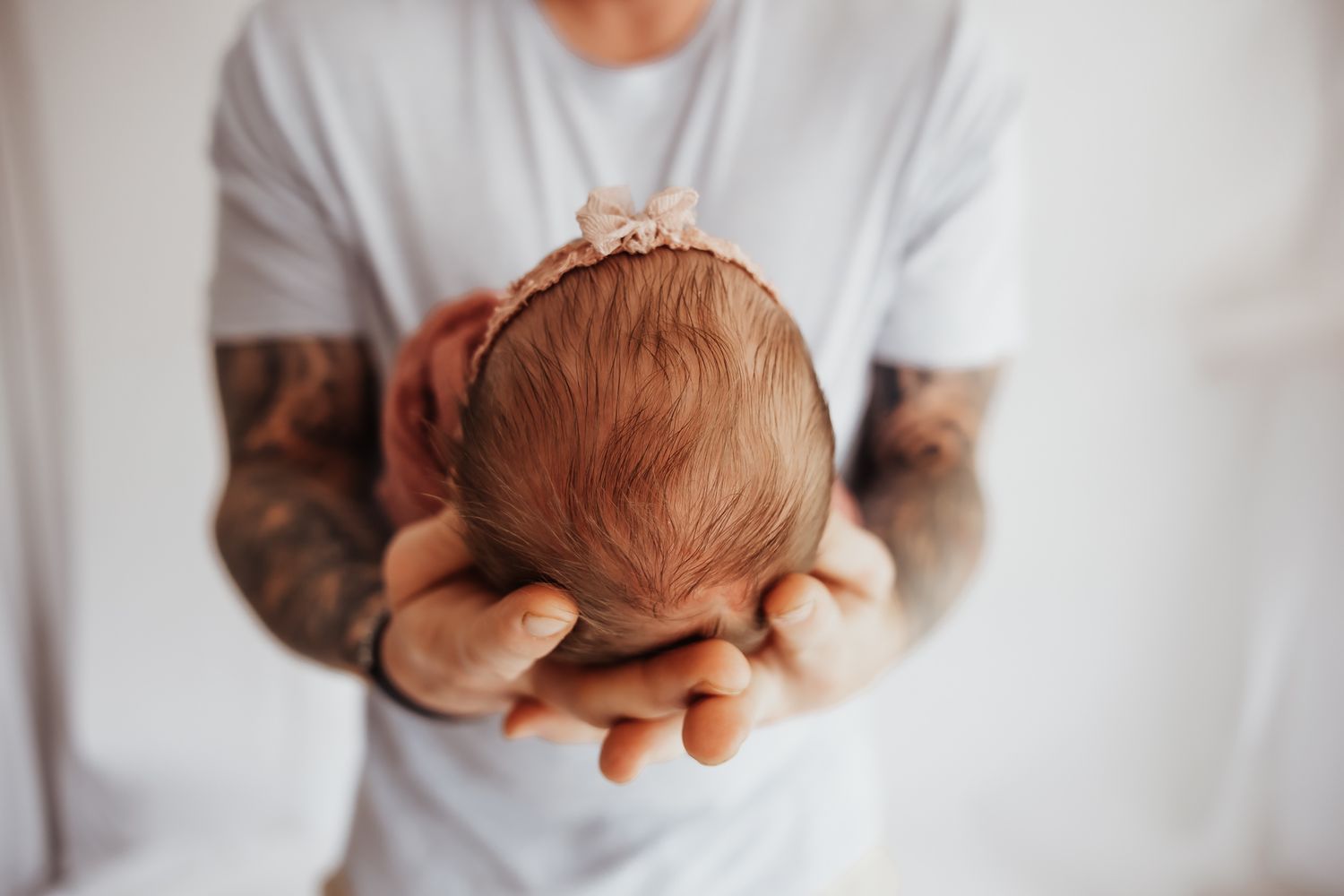 Tattooed father gently cradling sleeping newborn baby in both hands, close-up portrait of tender parental bond.