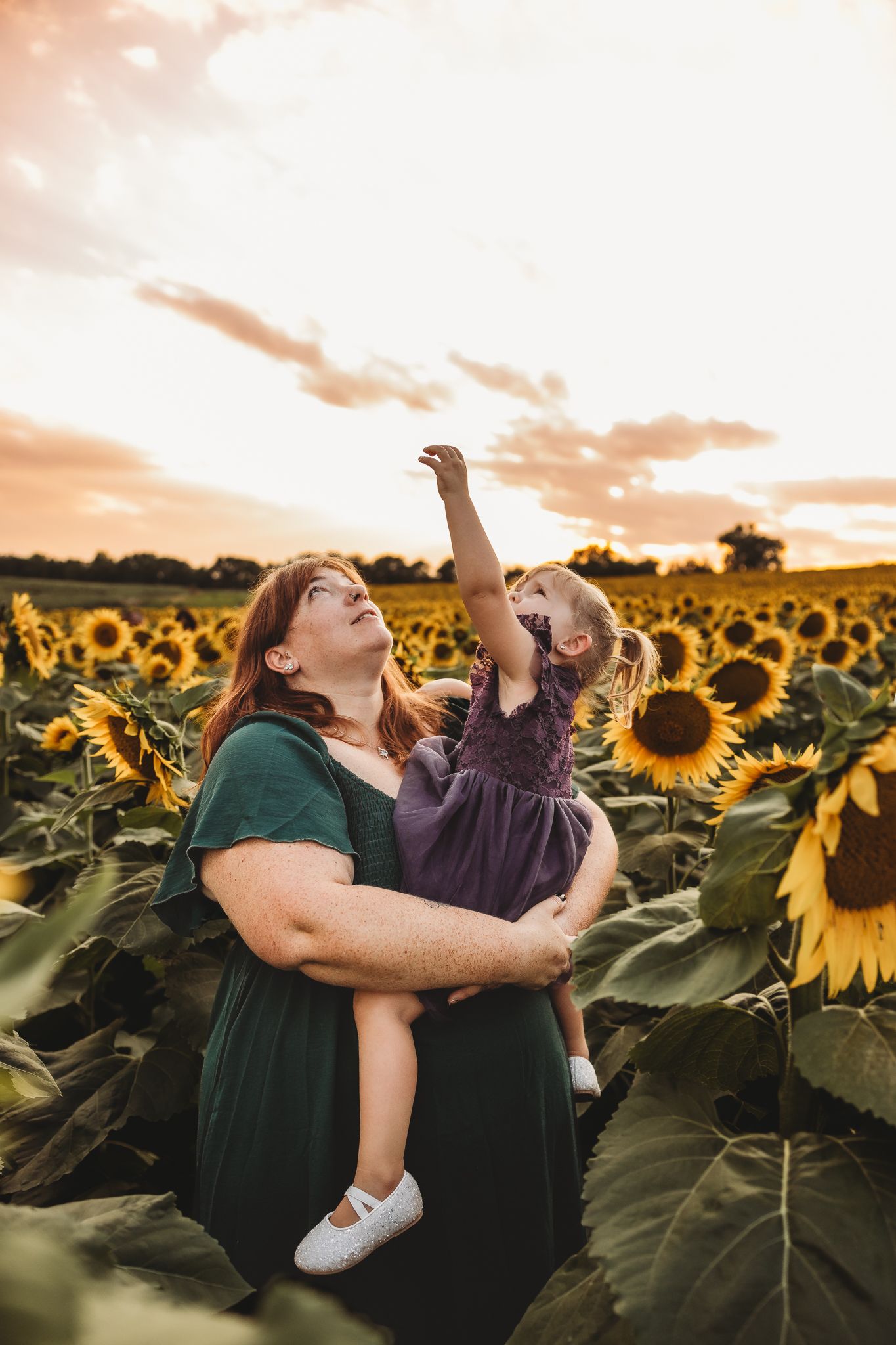 Mother holding toddler daughter in a sunflower field at sunset, child reaching up toward the sky.
