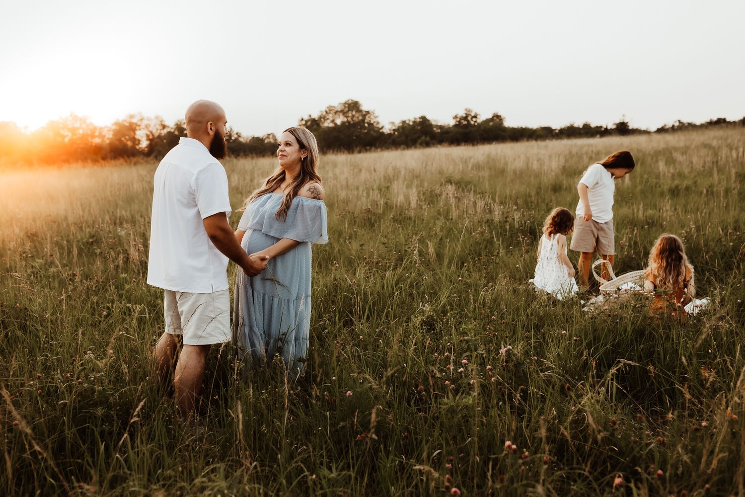 Family photo session in golden hour sunlit meadow with couple holding hands and children playing in background around Topeka, KS.