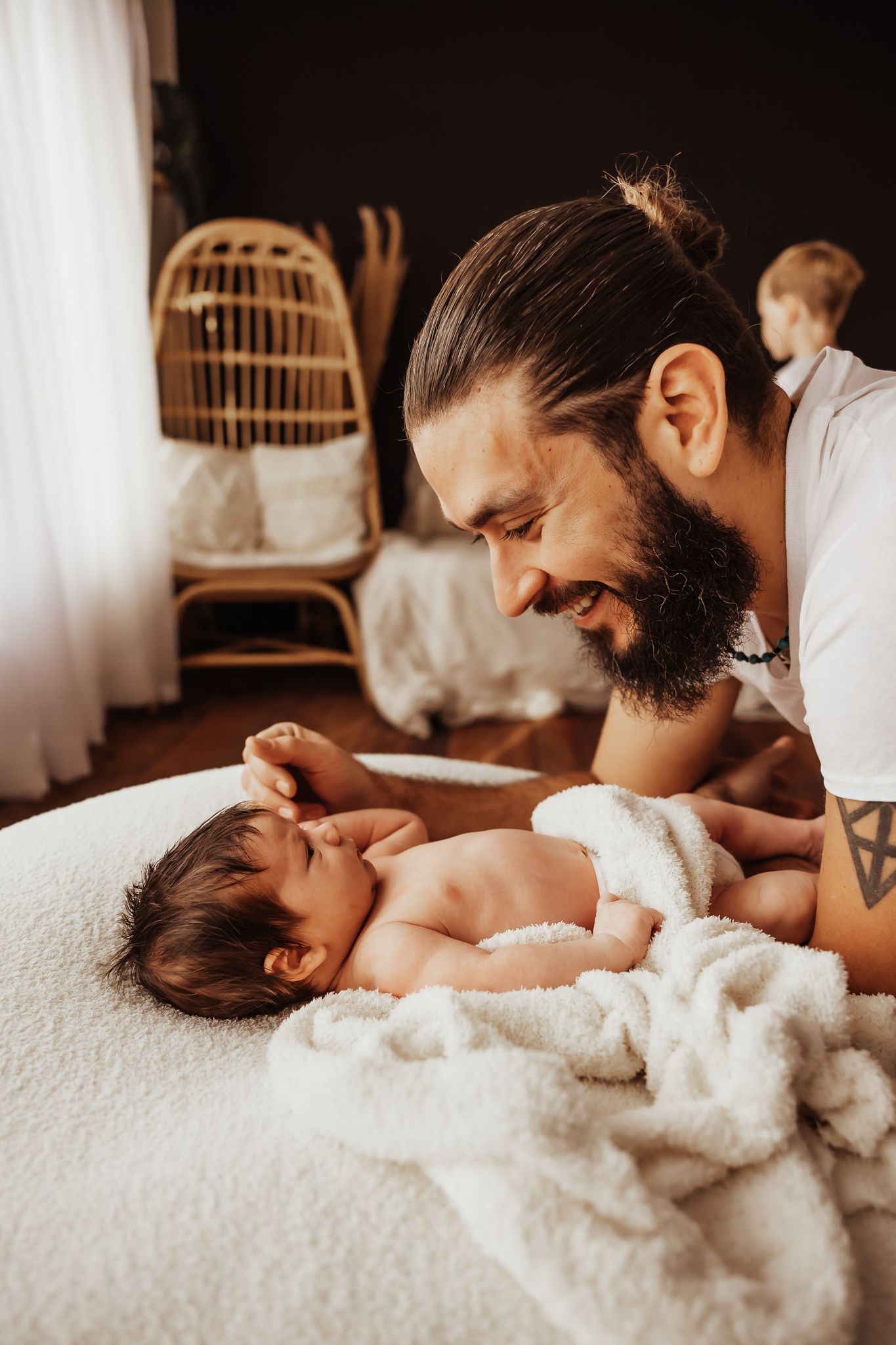 Bearded father smiling at his newborn baby lying on a white blanket on a bed in a cozy bedroom.