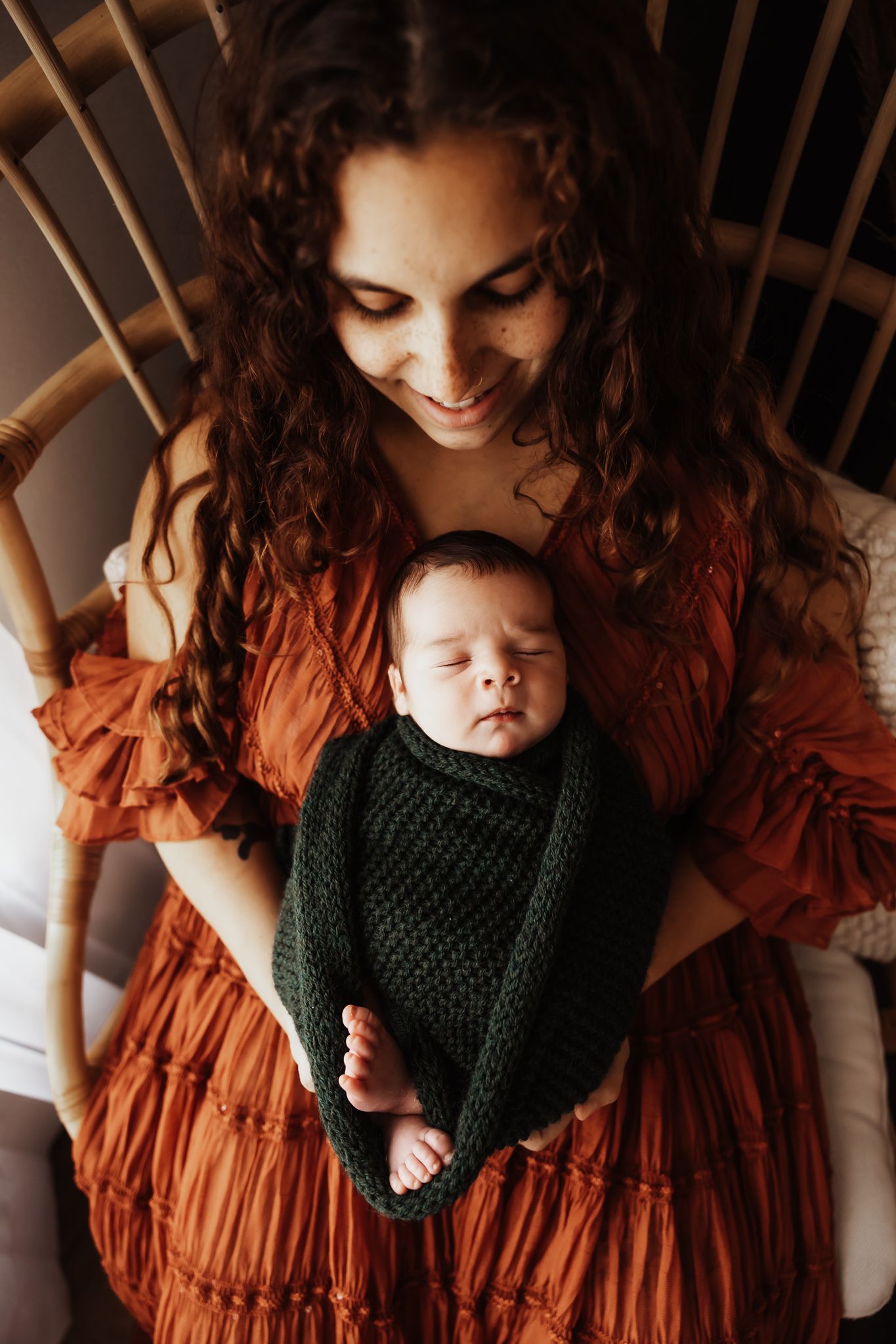 Smiling mother with curly red hair holds sleeping newborn wrapped in green knit swaddle while seated in rattan chair.