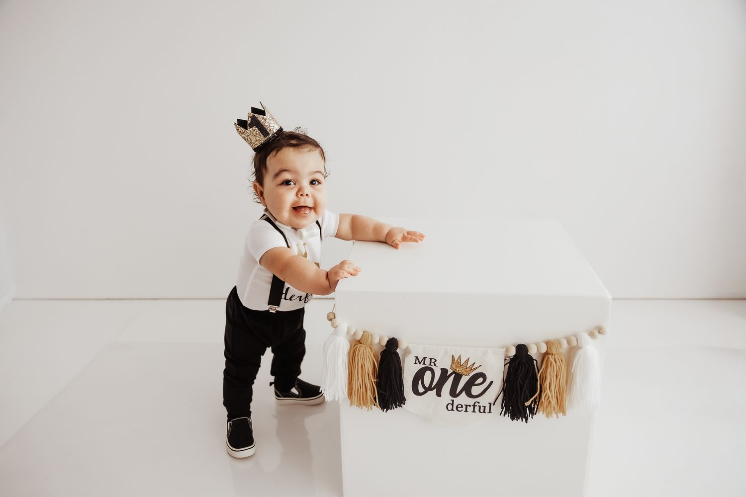 A baby boy in suspenders stands by a white cake smash table decorated with a “one”erful banner.