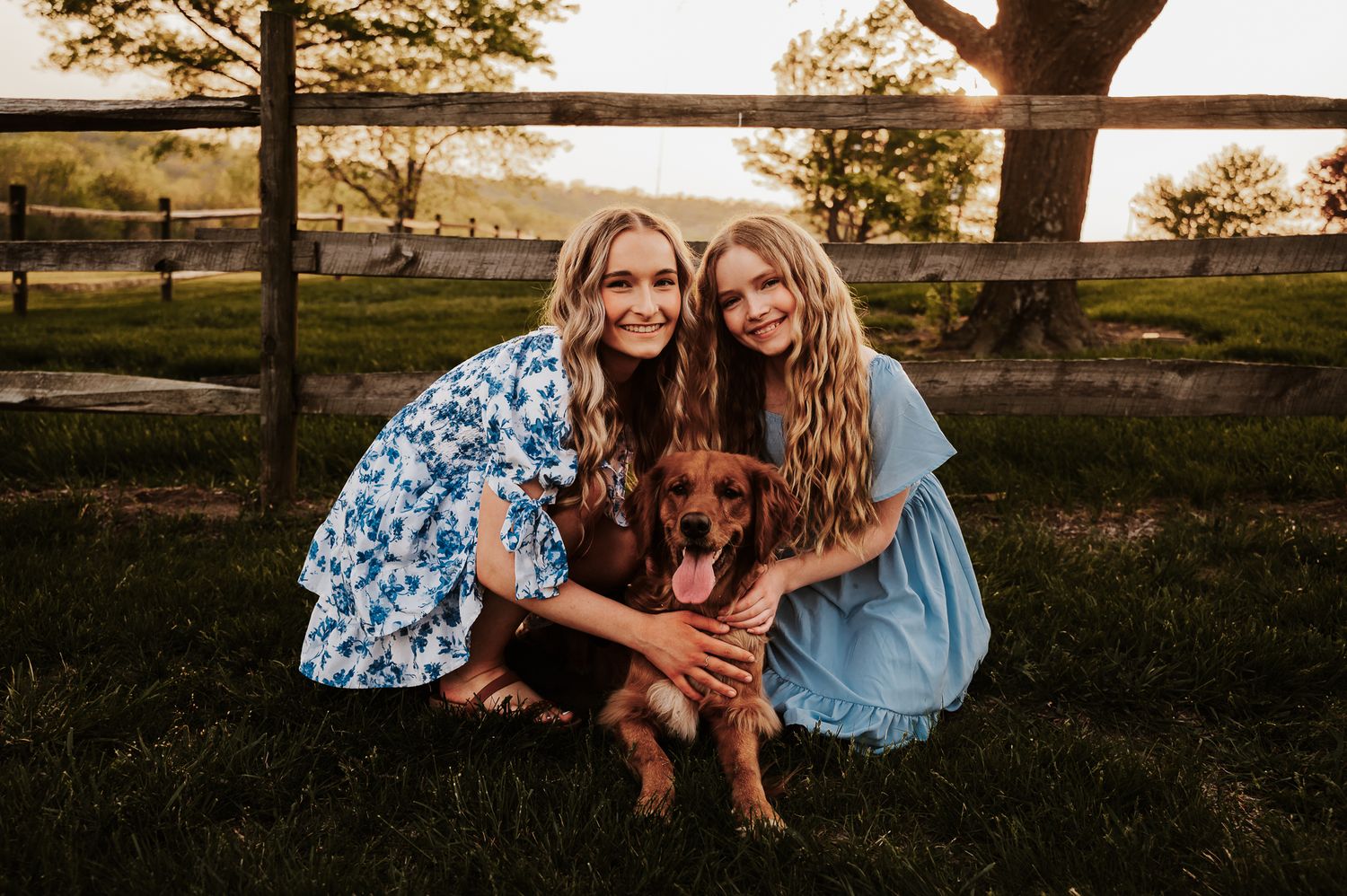 Two smiling girls in blue dresses crouch on green grass with a brown dachshund near a wooden fence at sunset.