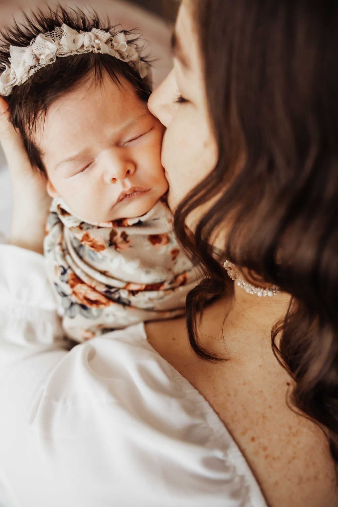 A mother gently kisses her sleeping newborn baby wrapped in a floral swaddle blanket wearing a delicate flower headband.