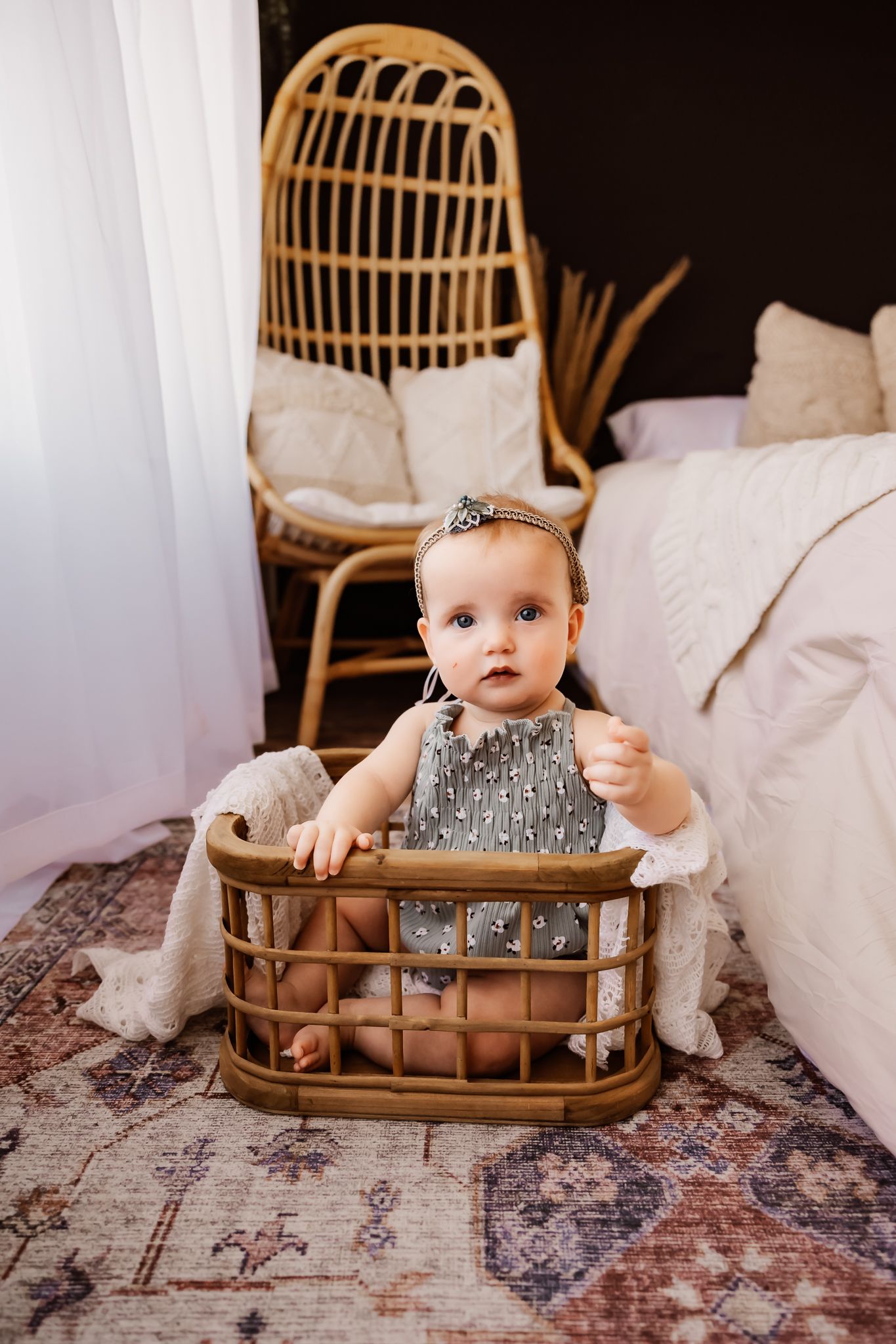 Adorable baby girl in gray dress sitting in wicker basket on boho rug with rattan chair in background.