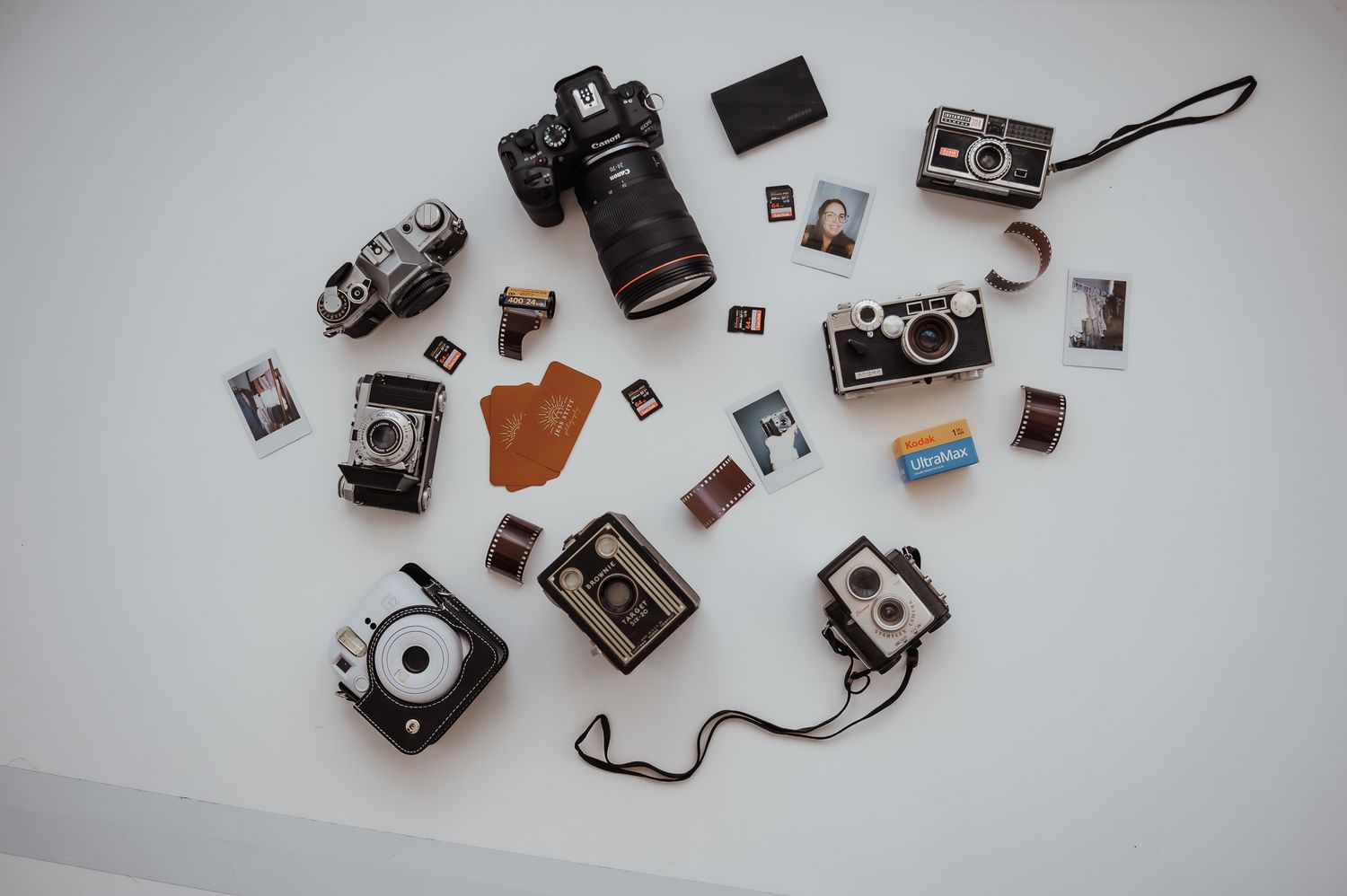 Flat lay collection of vintage and modern cameras with film and memory cards on white surface.