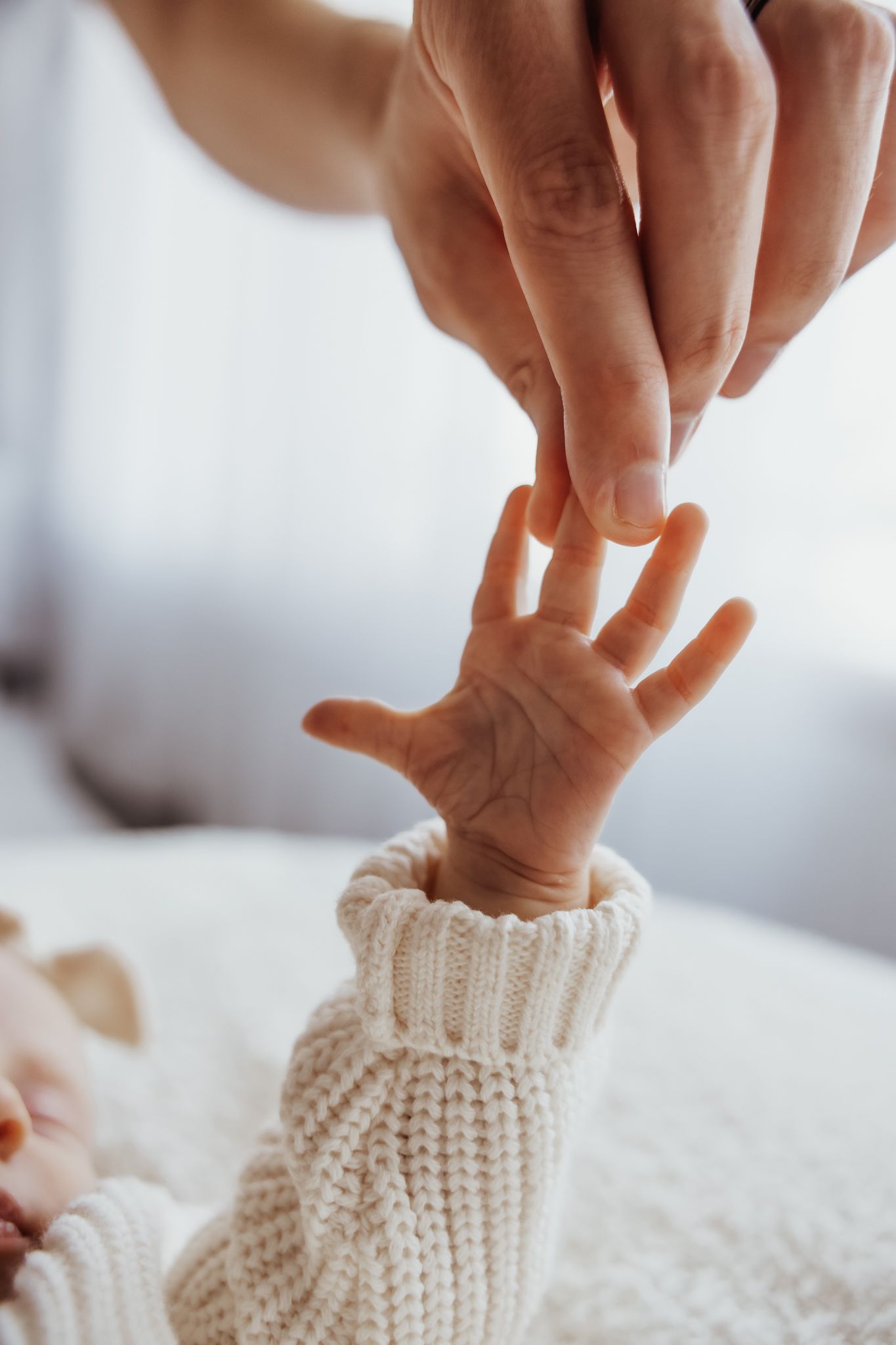 Close-up of an adult hand gently holding a tiny baby hand wearing a white knit sweater.