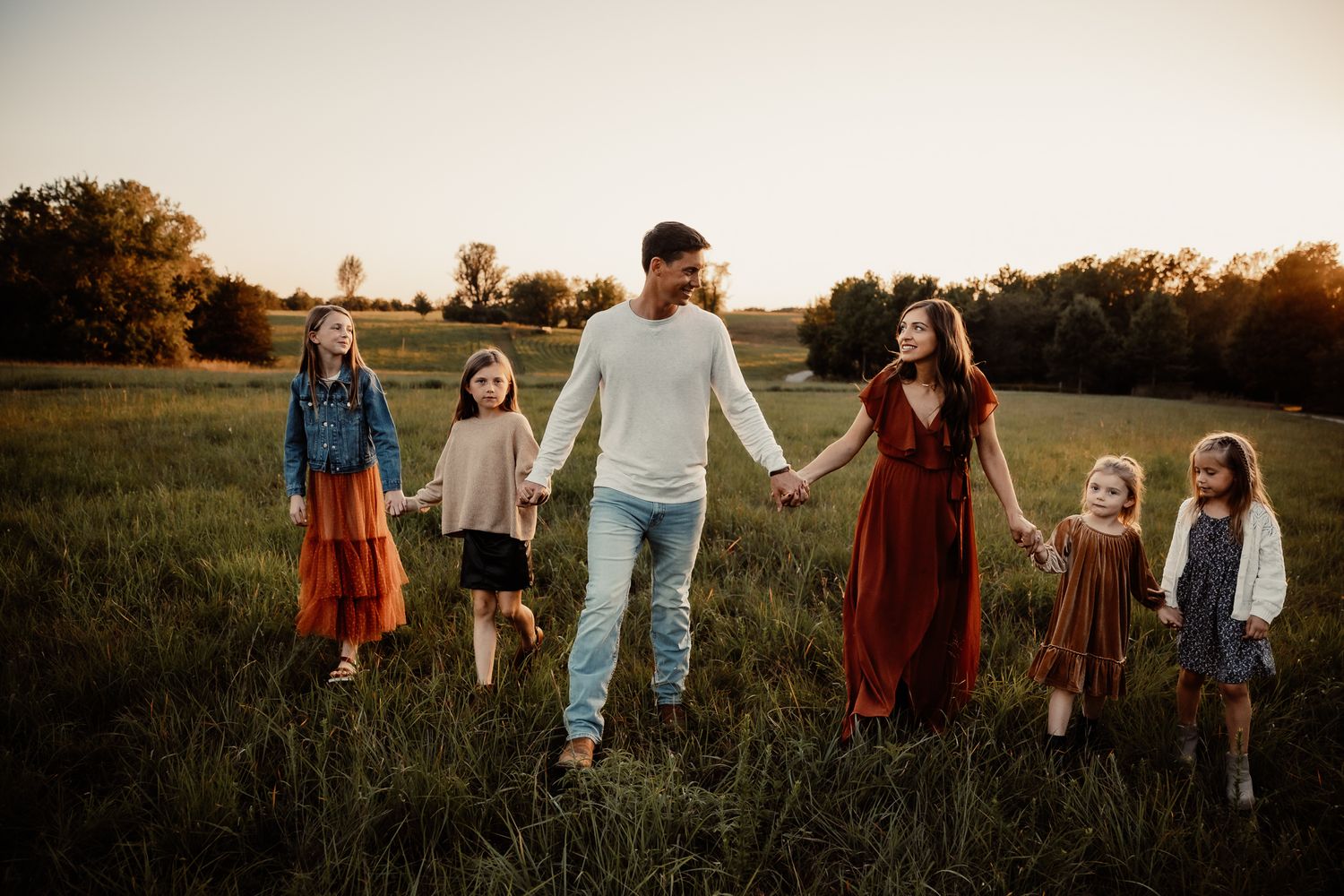 Family of six walking hand-in-hand through a sunlit green field at golden hour in warm autumn tones.
