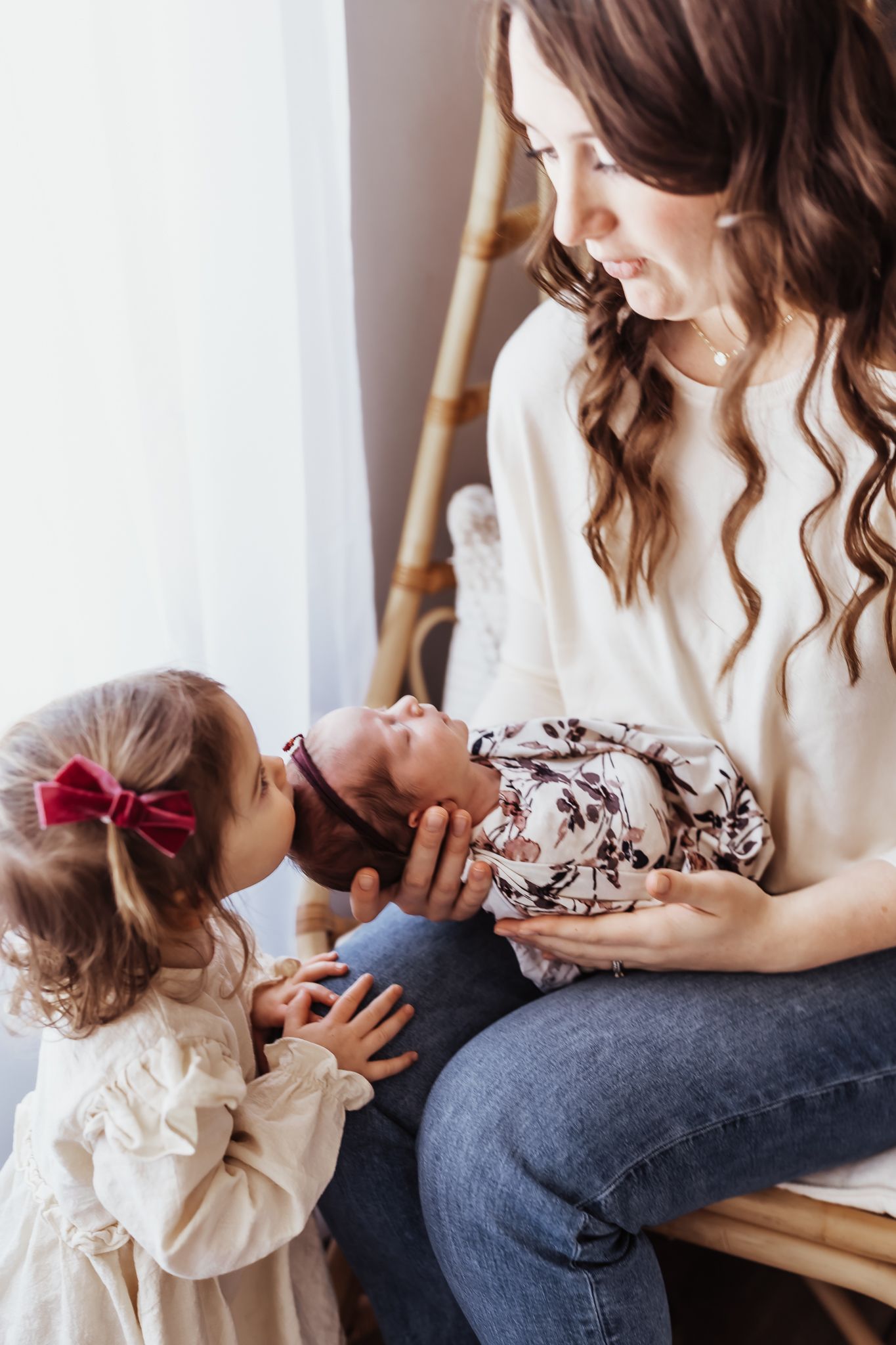 A mother with curly brown hair holds her newborn while a toddler girl in a white dress kisses the baby.