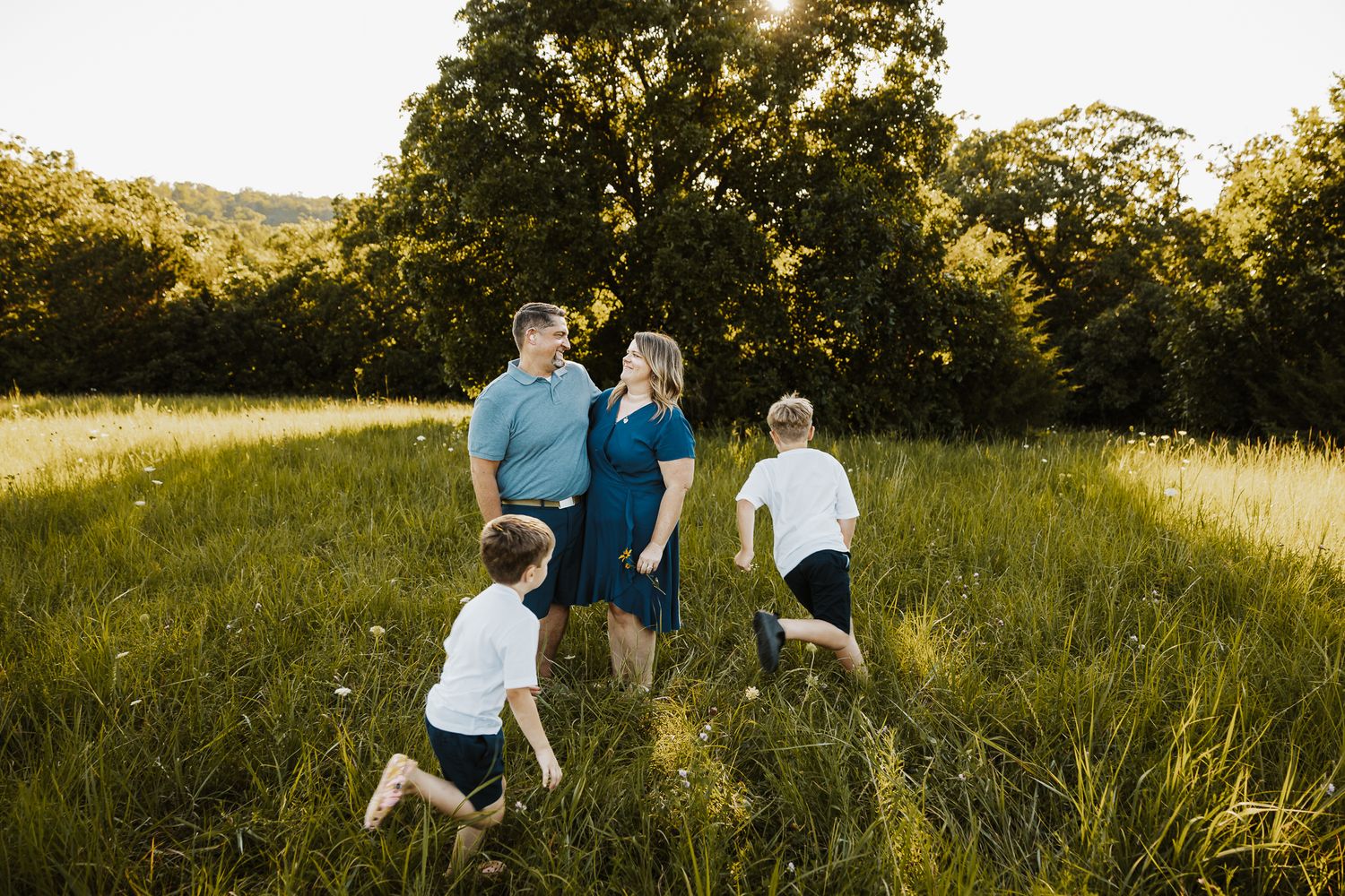 Family of four enjoying a sunny outdoor photo session in a lush green meadow surrounded by trees.