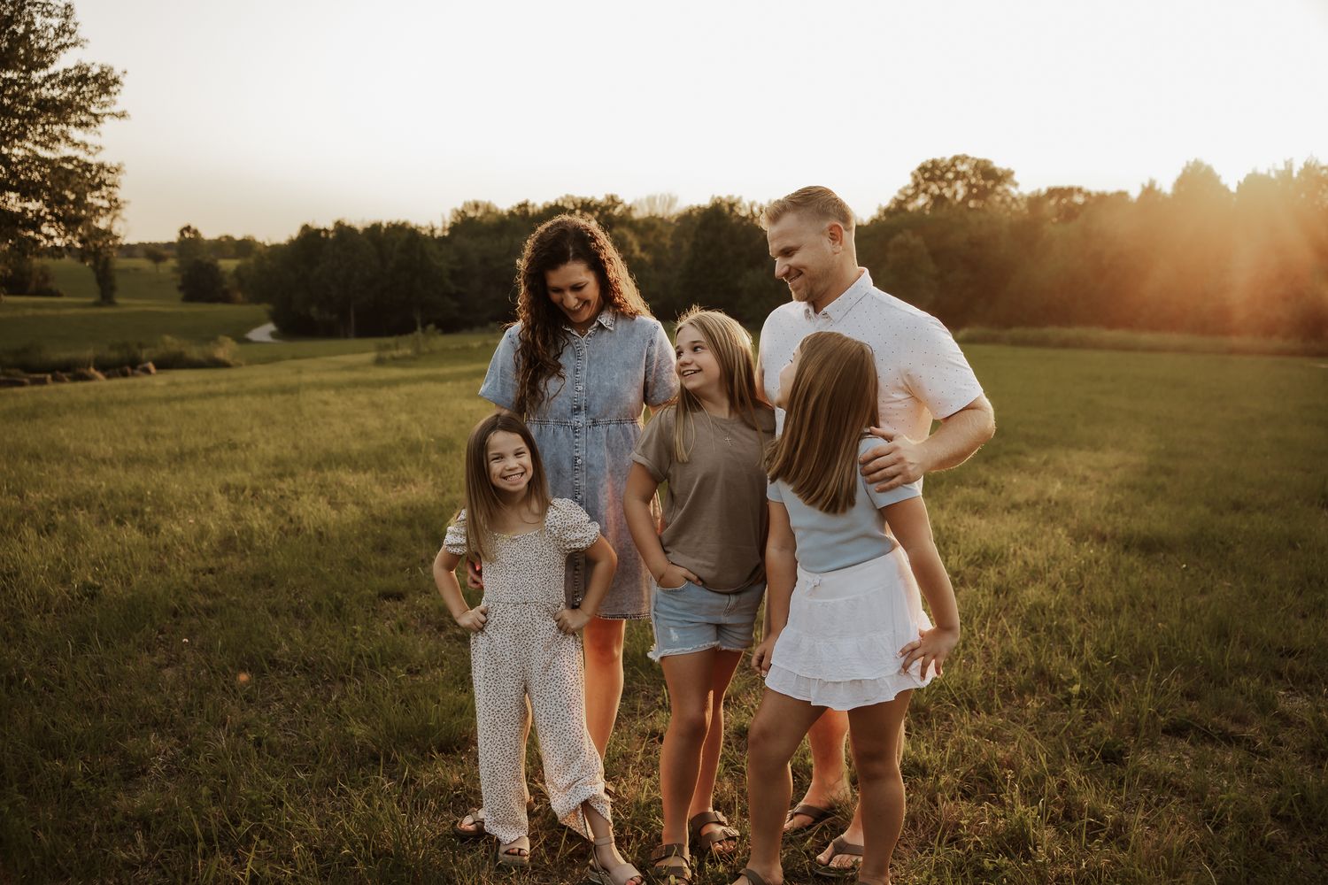 Family of five laughing together in a sunlit green field during golden hour sunset outdoors.