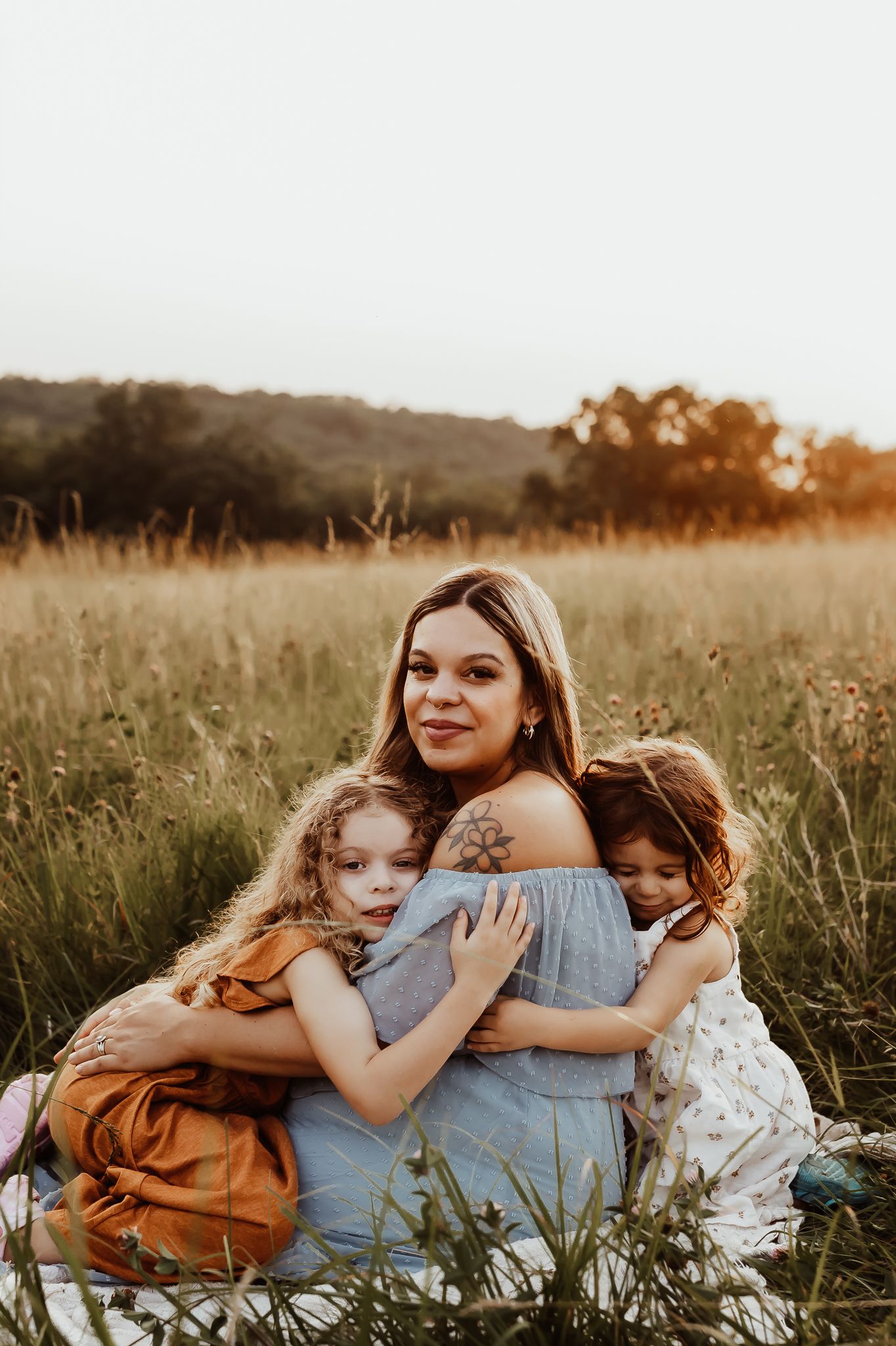 A mother sits in a golden field at sunset, warmly embracing her two young daughters.