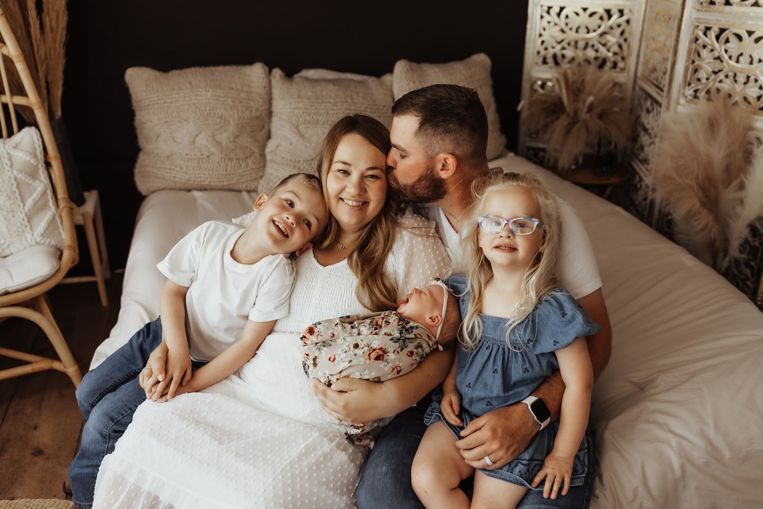 Happy family of five sitting on a bed, parents kissing with three children including a newborn baby in a cozy home setting.