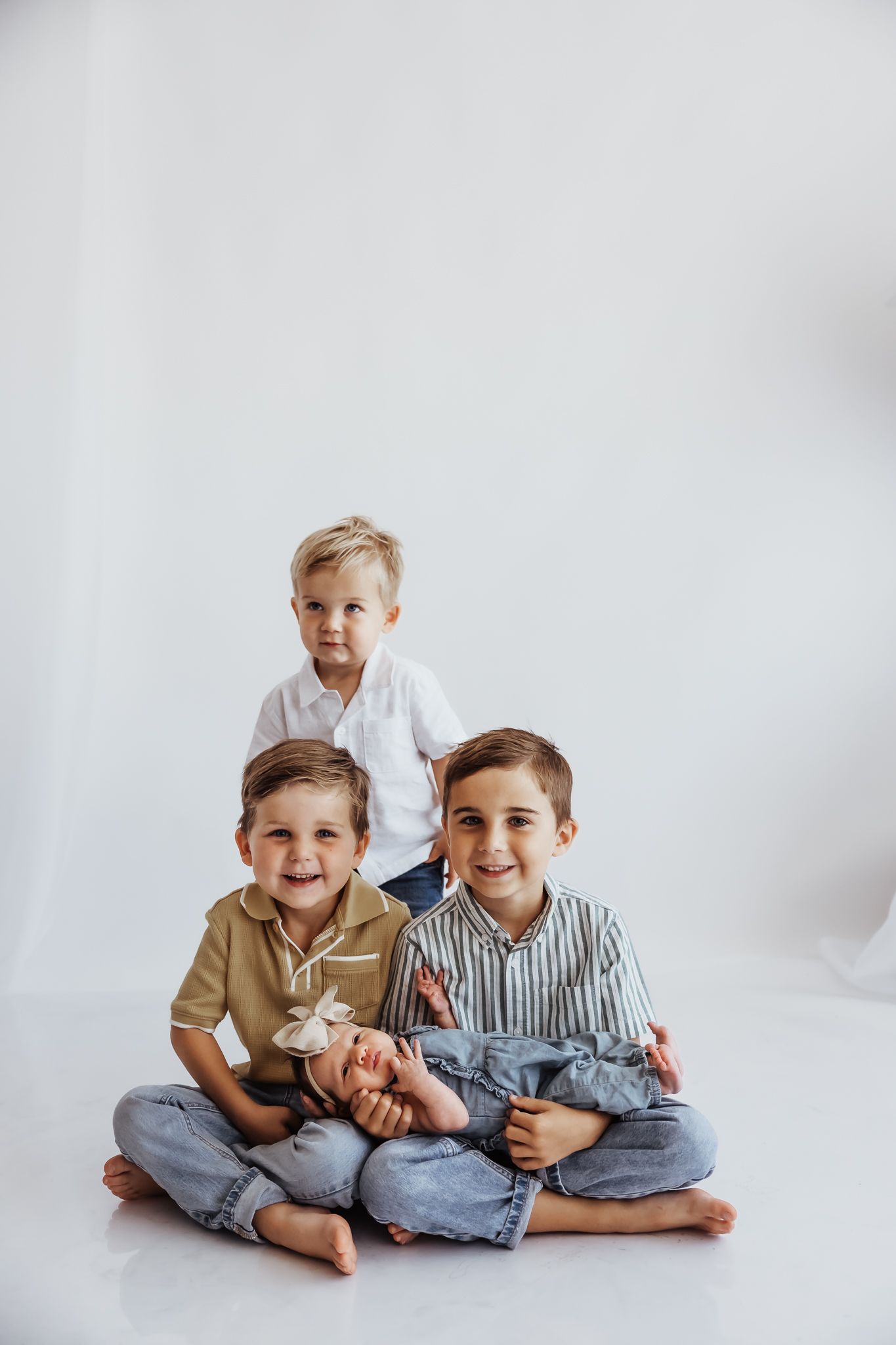 Three young boys sitting together on white background, two cross-legged holding a baby, one standing behind, smiling.