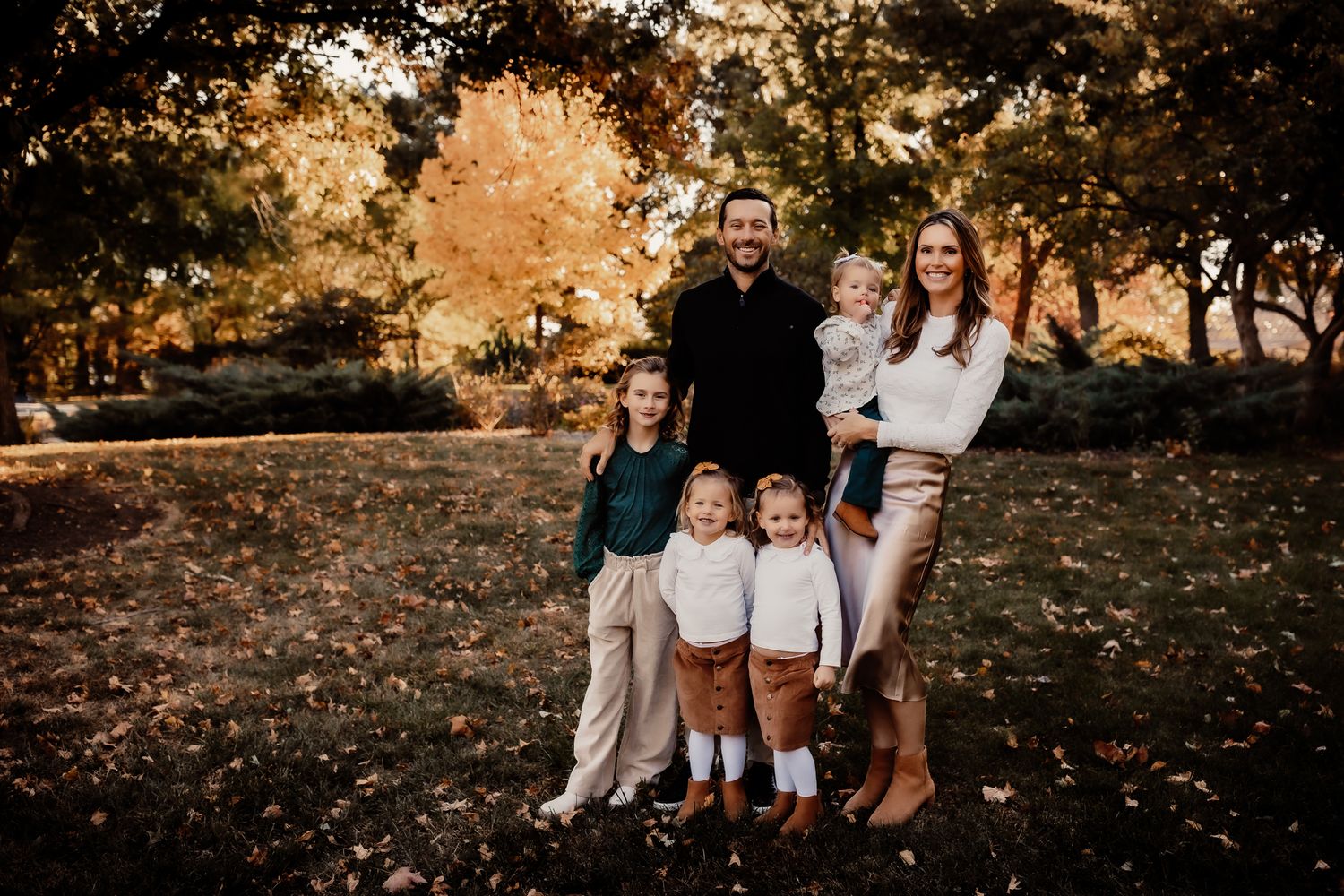 Family of six posing outdoors in autumn park with colorful fall foliage and green grass background.