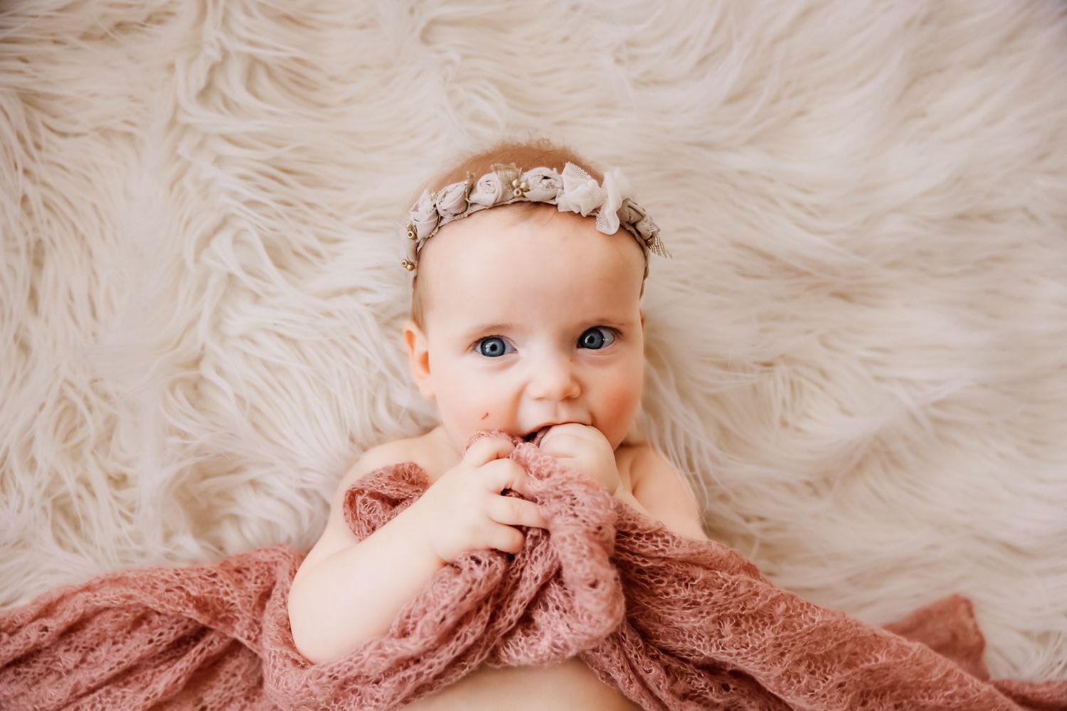 Adorable baby girl with blue eyes wearing a floral headband, wrapped in a pink blanket on a fluffy white fur rug.