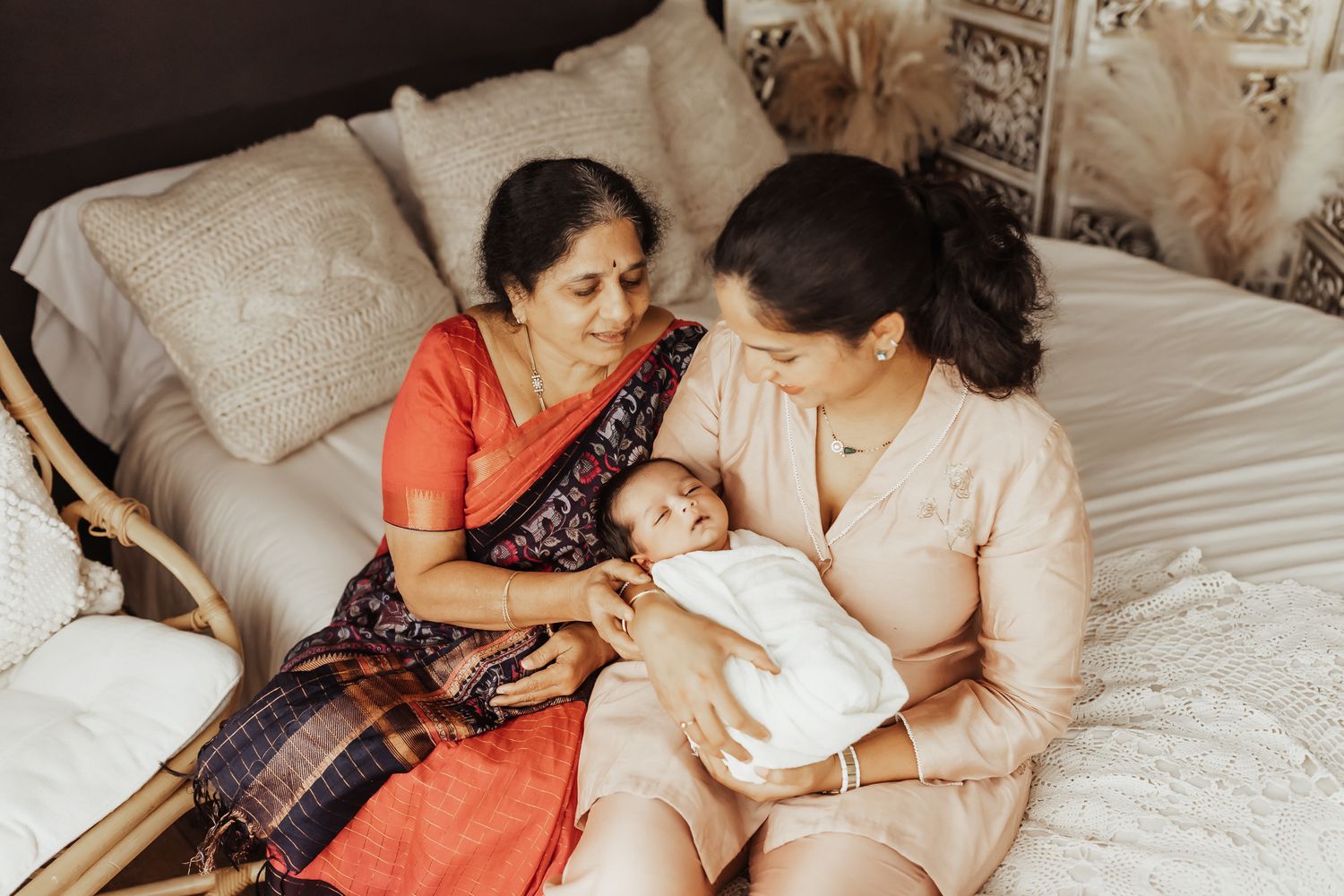 Two women in traditional Indian attire sit on a bed, lovingly cradling a newborn baby wrapped in white.
