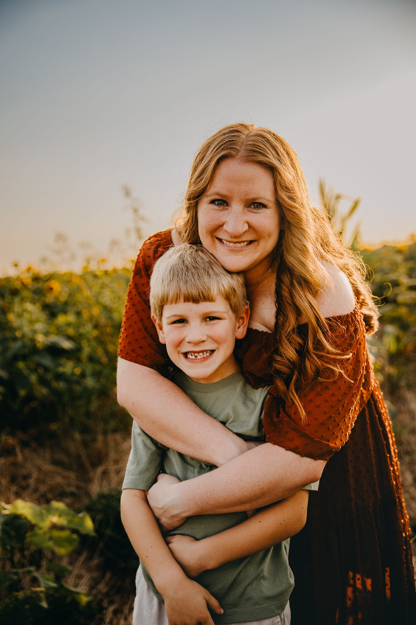 A smiling mother hugs her young red-haired son in a sunflower field at sunset.
