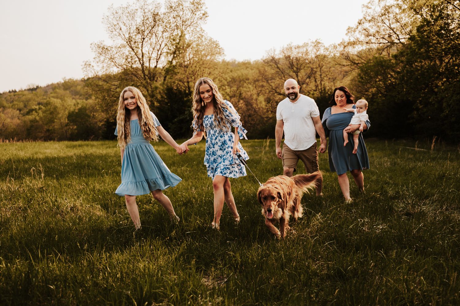 Family with two girls, a baby, and a golden retriever dog walking through a sunny green meadow at golden hour.