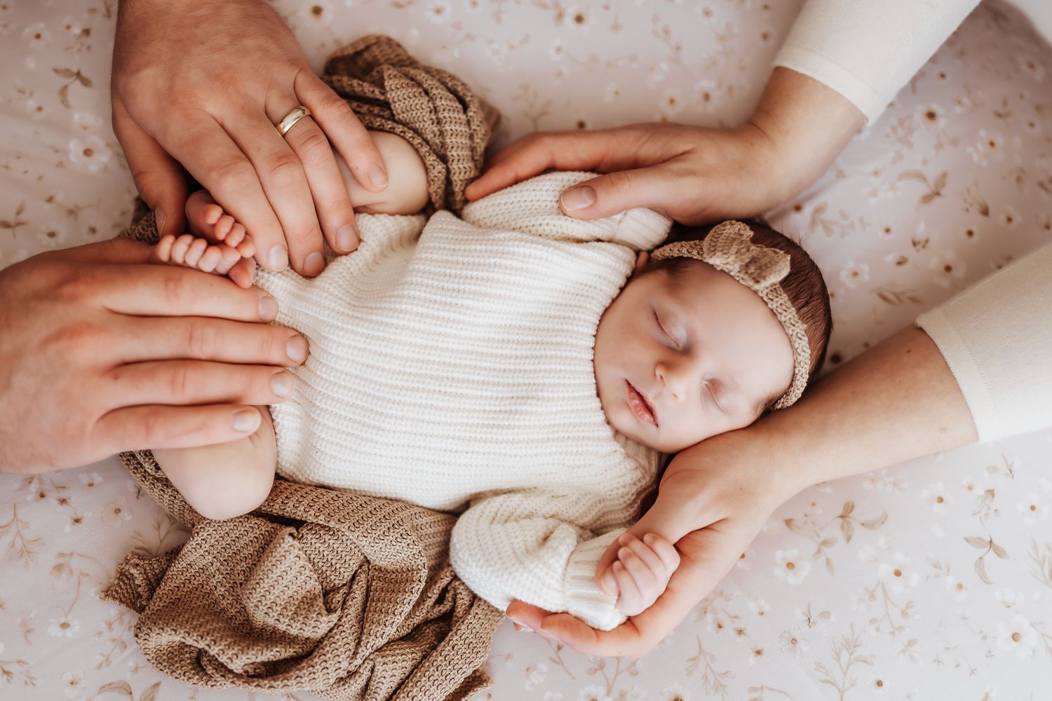 Sleeping newborn baby in white onesie with bow headband, cradled by parents hands on a soft floral blanket.
