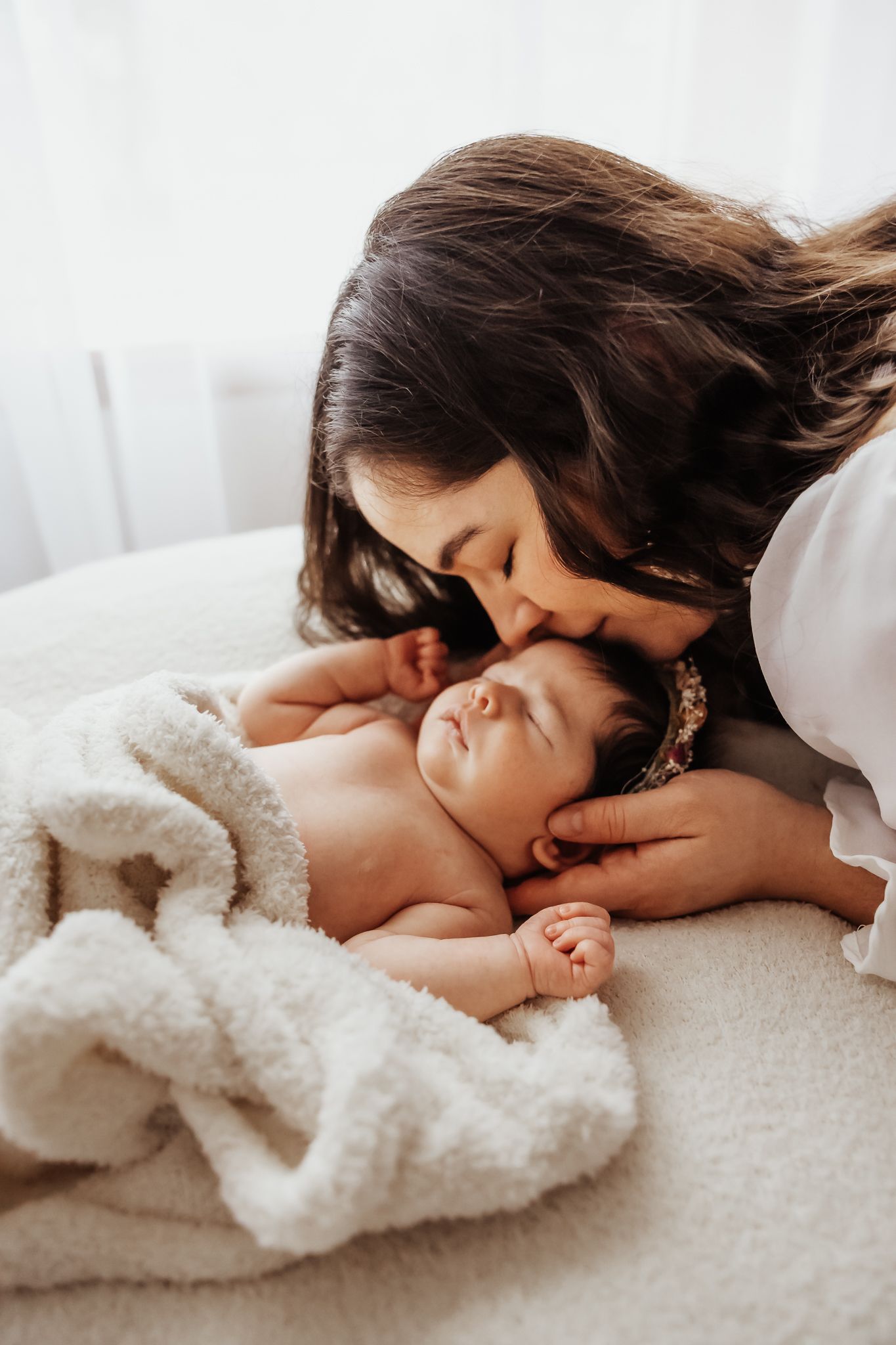 A mother with dark hair tenderly kisses her sleeping newborn baby wrapped in a soft white blanket.