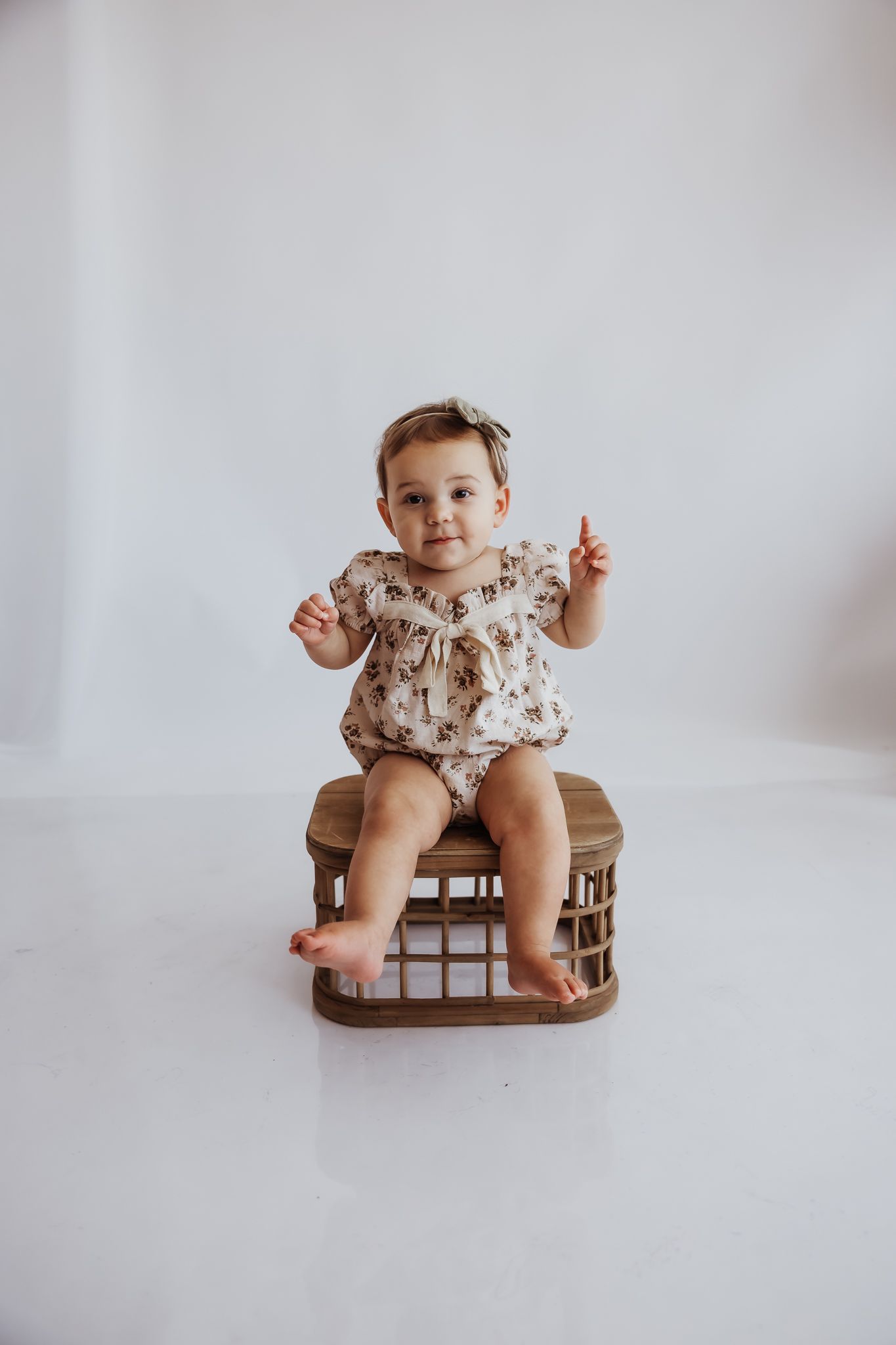 A cute baby girl in a floral romper and bow headband sits on a wicker box, raising one finger, against a white background.