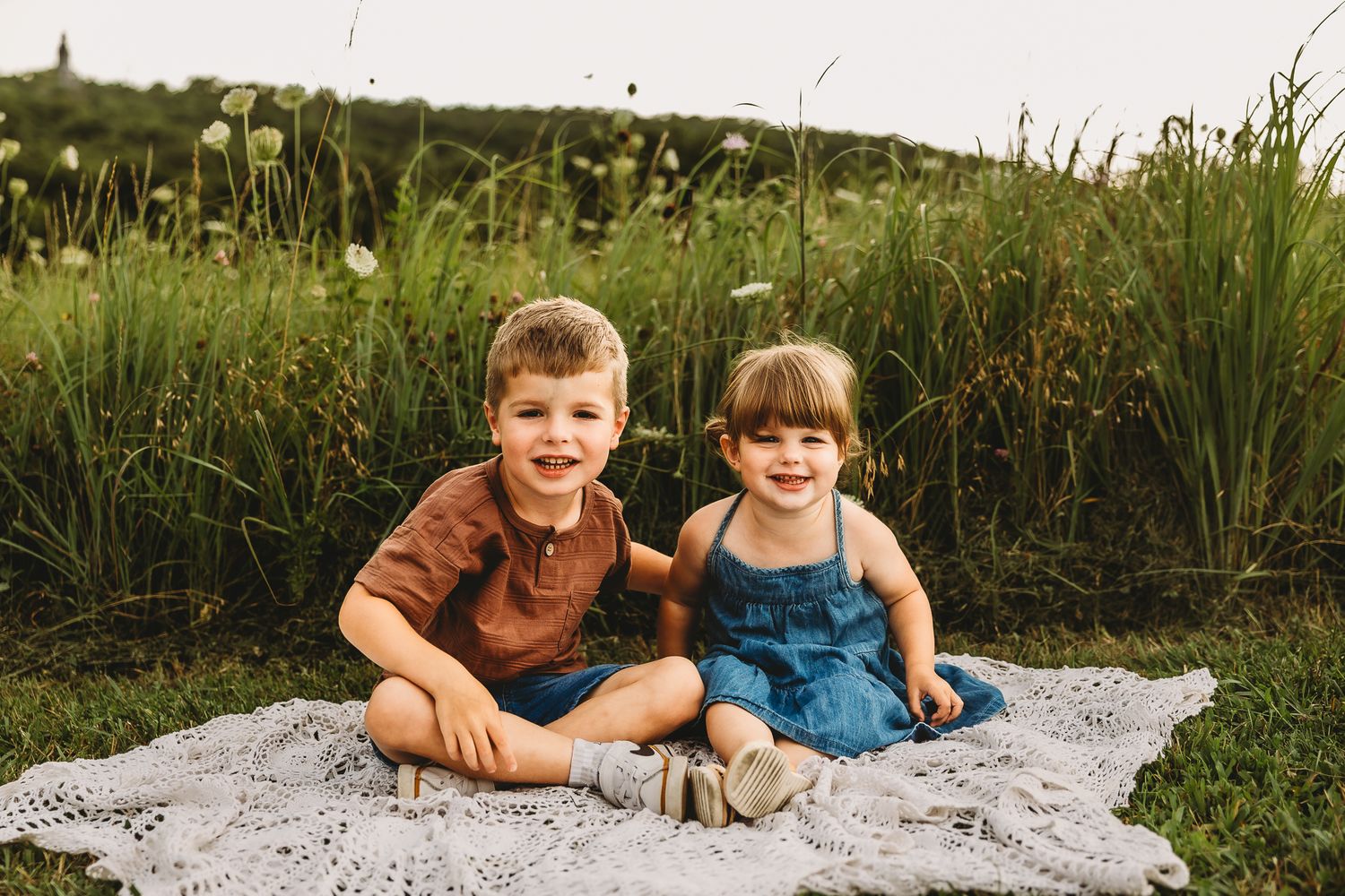 Adorable young brother and sister smiling together on crochet blanket in green summer field.